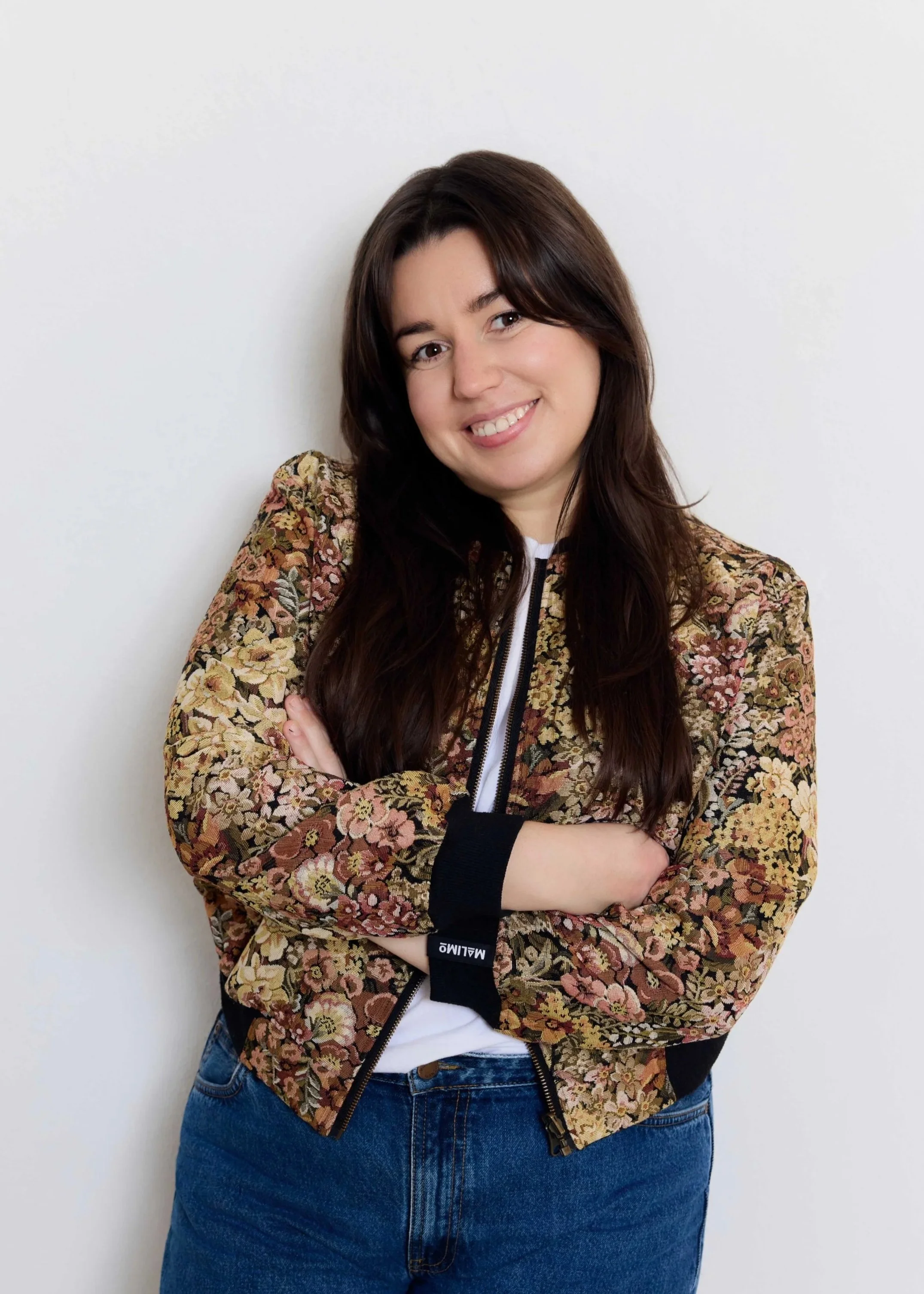 A young woman with long dark hair wearing a patterned bomber jacket and blue jeans, standing against a white wall, smiling with arms crossed.