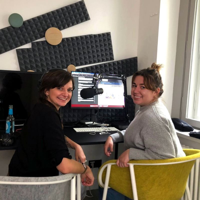 Two women sitting at a desk with microphones and a computer screen in a recording studio, smiling at the camera.