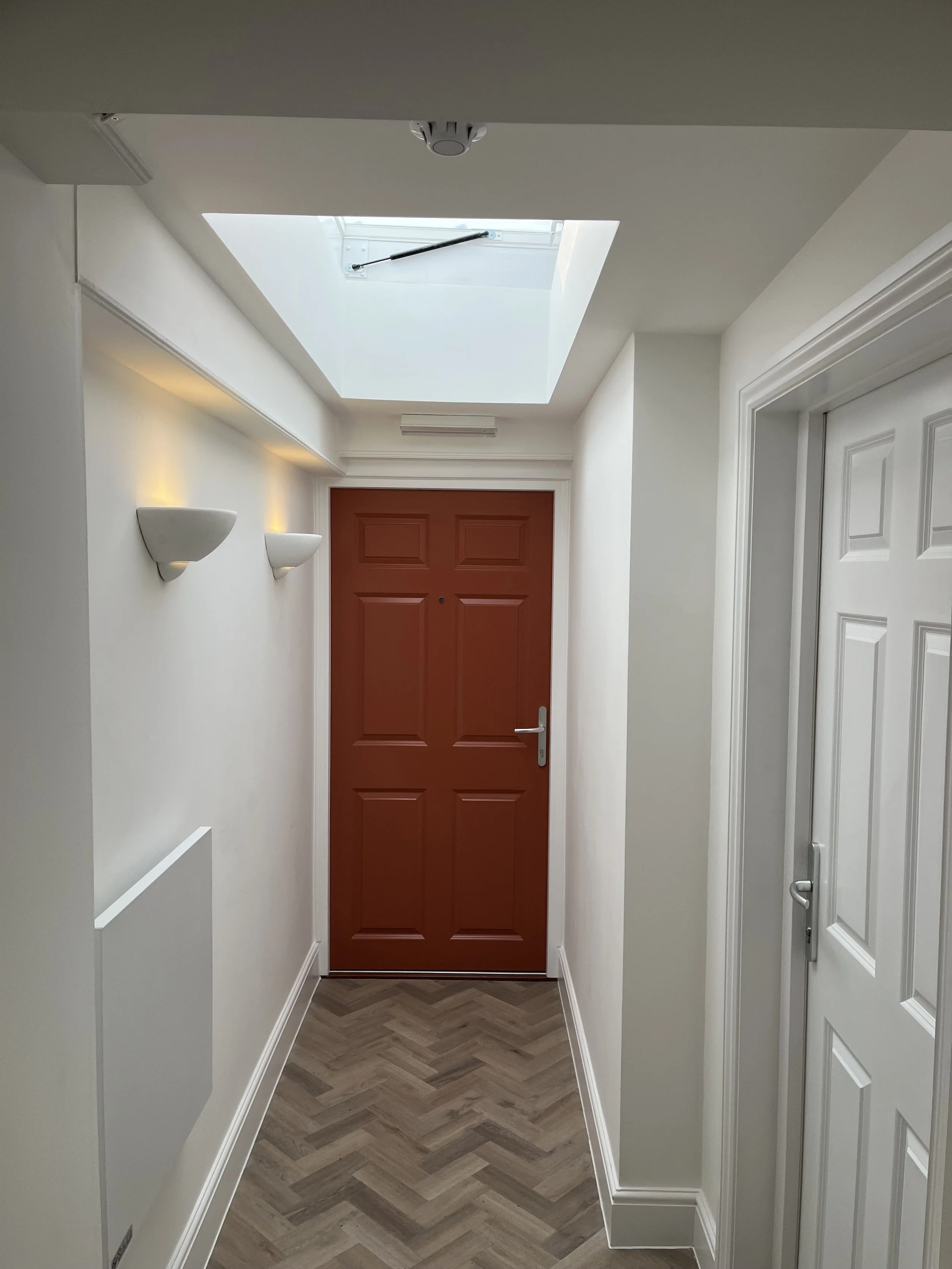 A corridor with white walls, a red front door at the end, white wall-mounted lights, a skylight in the ceiling, and hardwood herringbone floor.