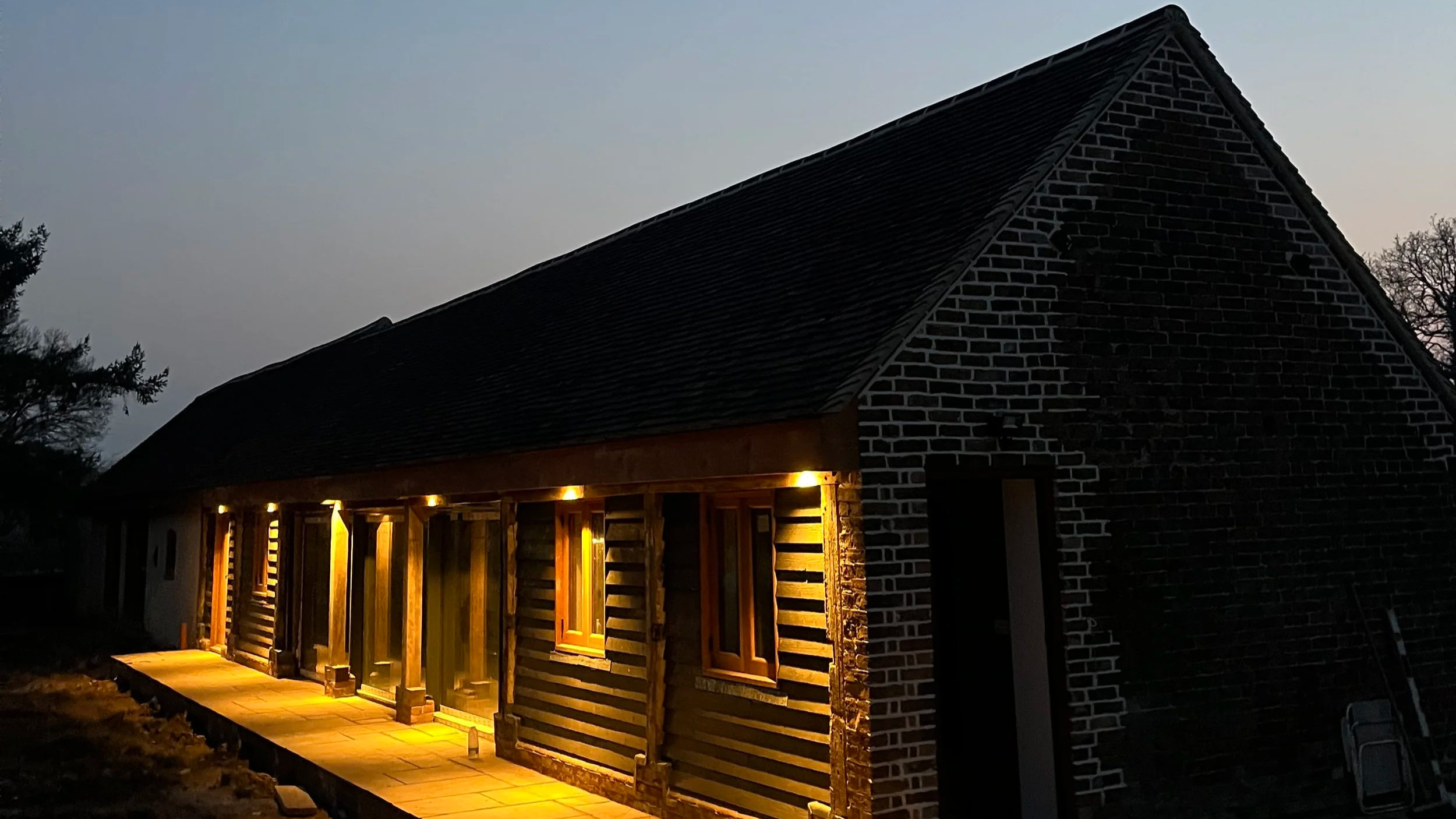 A house with brick walls and a steep roof, illuminated by exterior lights, at dusk or early evening.