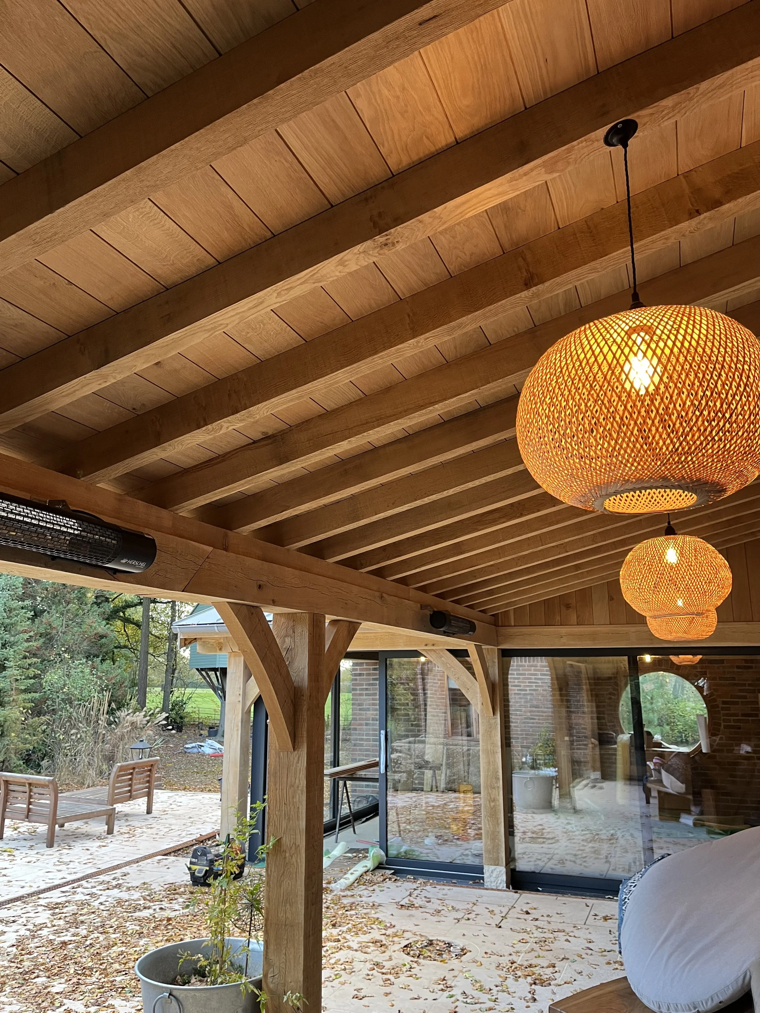 A wooden outdoor patio with a ceiling made of wooden planks, decorated with hanging woven orange lantern-style lights. The patio is attached to a house with large glass doors, and there are outdoor chairs and a table visible in the yard.
