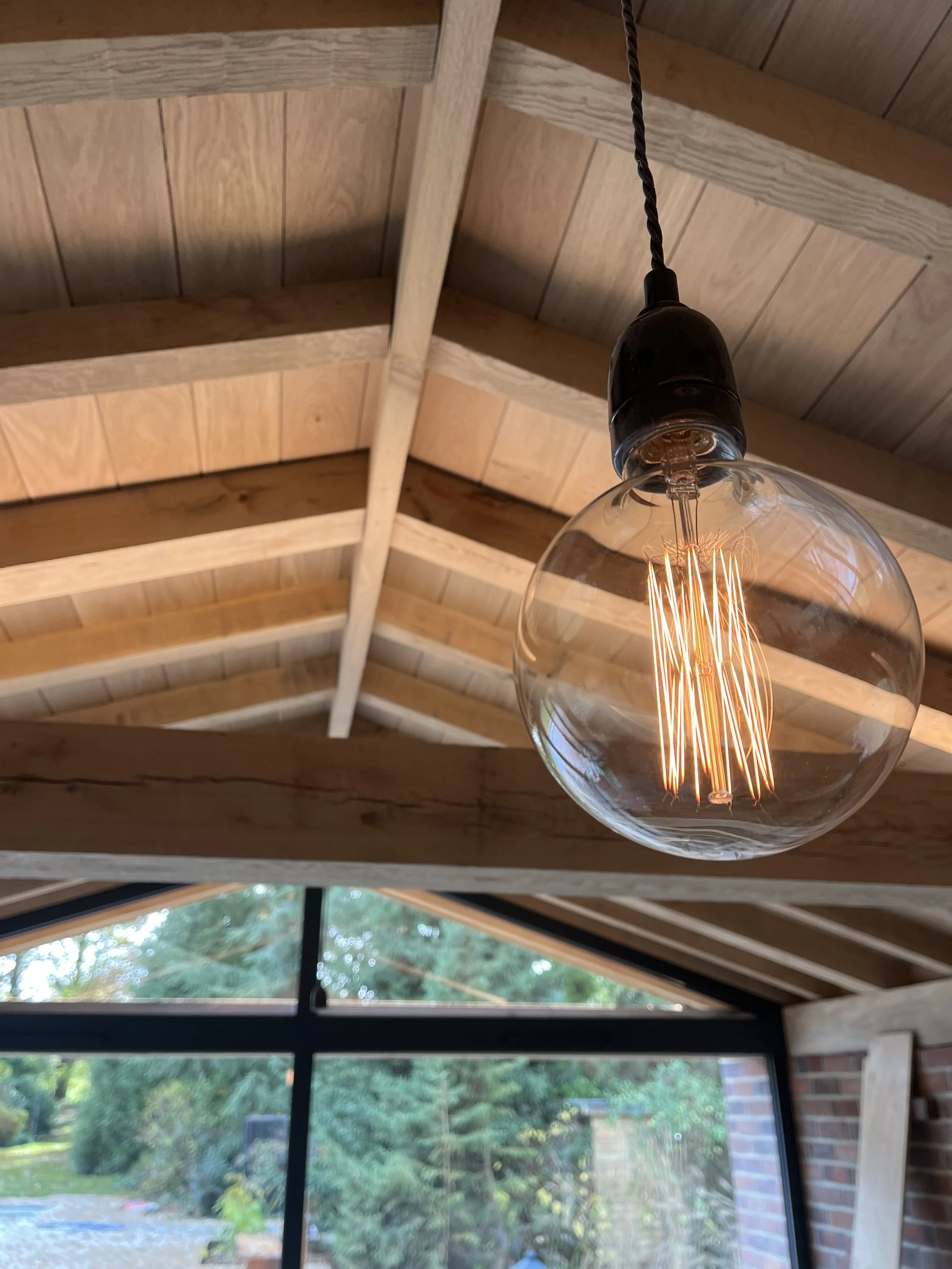 Close-up of a black pendant light with a glowing filament hanging from a wooden ceiling with beams, overlooking a green garden outside.