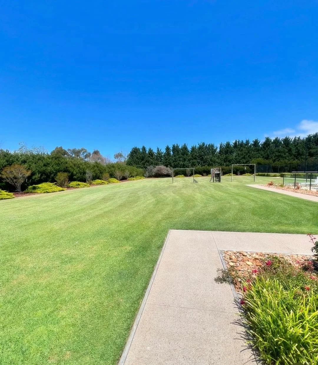 View of a well-maintained grassy park with a concrete pathway, landscaped bushes, and trees under a bright blue sky.