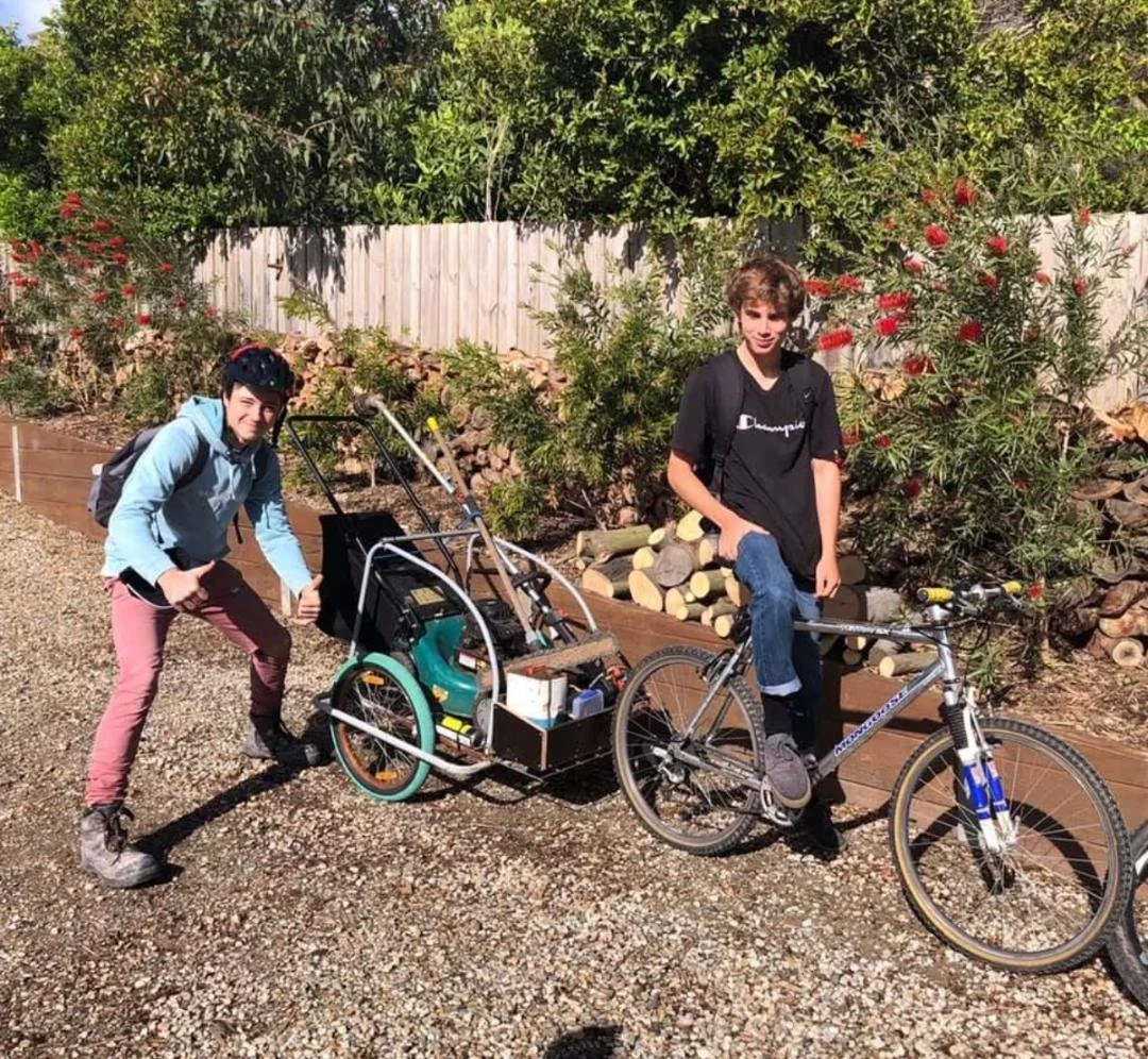 Two young men with backpacks posing with a bicycle and a bicycle trailer filled with tools and equipment on a gravel path, with green trees, bushes, and a wooden fence in the background.