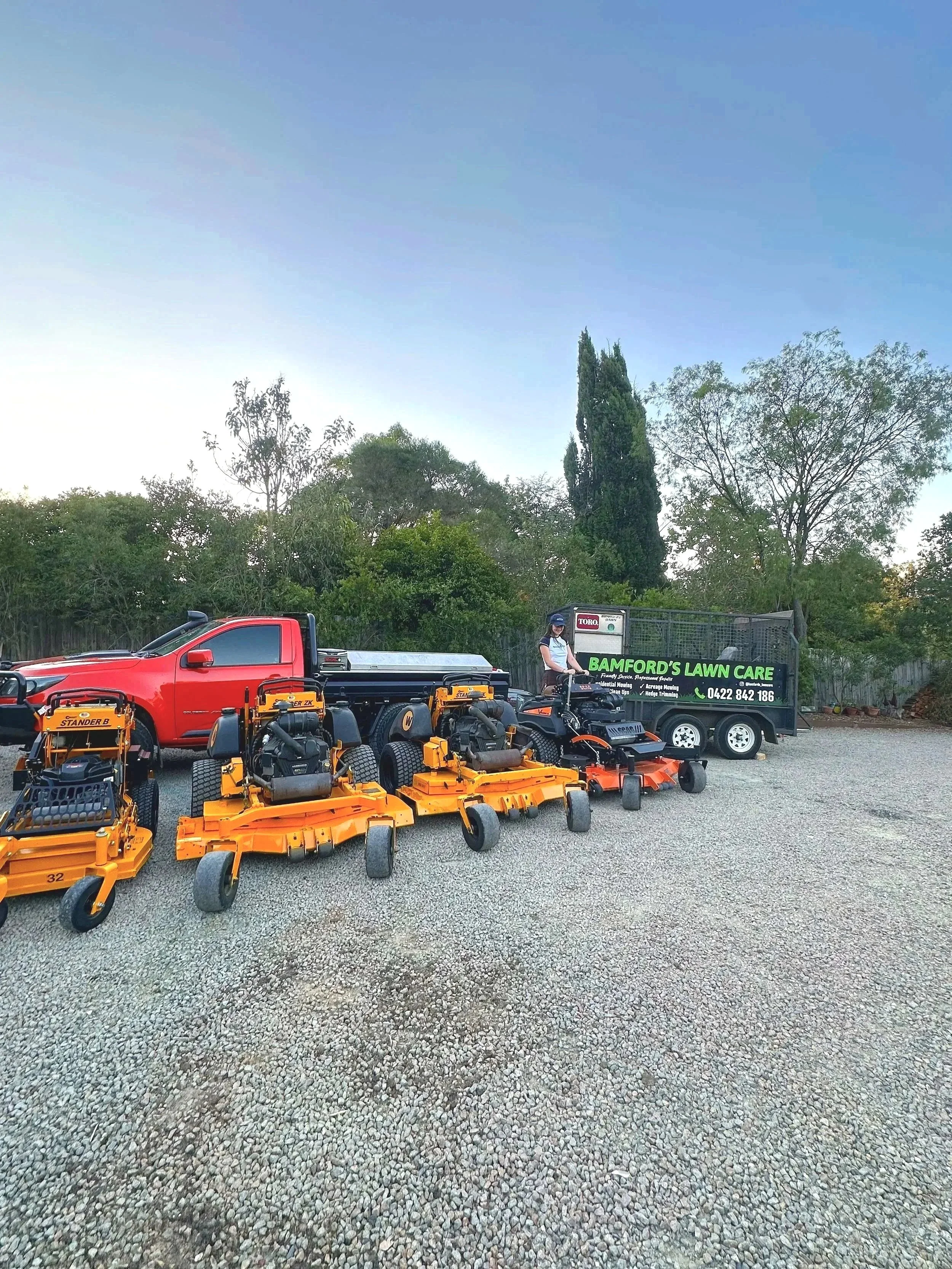 A collection of lawn mowing and landscaping equipment arranged in front of a parking lot containing a red pickup truck, with trees and a clear sky in the background. There is a green sign that reads 'Bamford's Lawn Care' with contact information.
