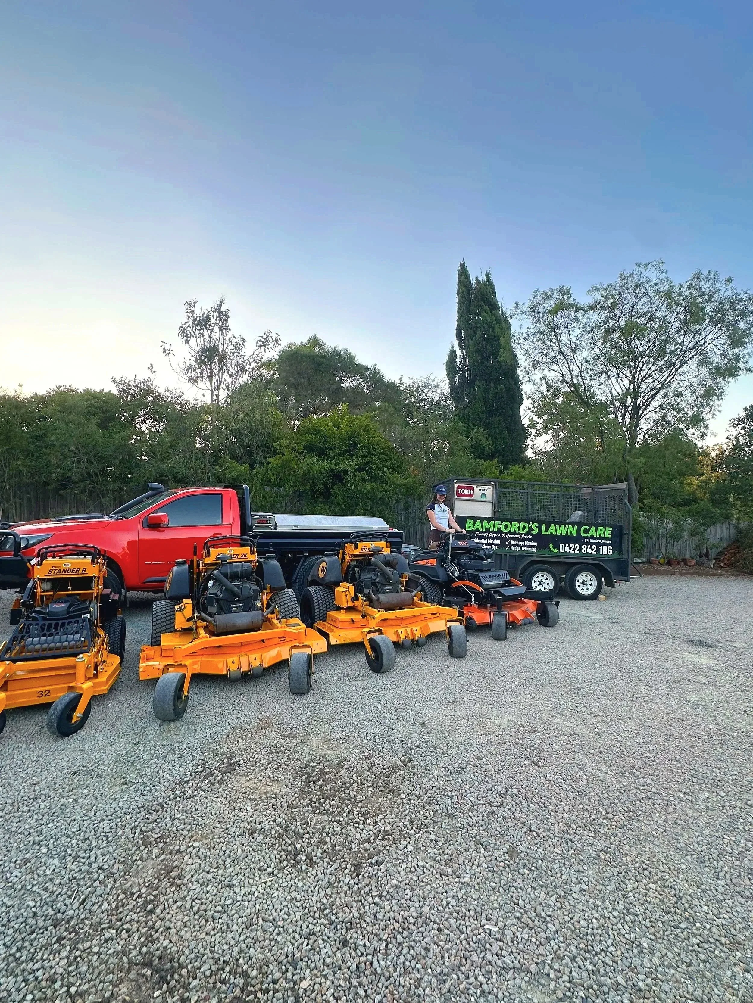 A row of four orange lawn mowers placed on a gravel area in front of a red truck and a green trailer with a sign that reads 'Bamford's Lawn Care' on it. A person in a uniform is standing on one of the lawn mowers. There are trees and a clear sky in the background.