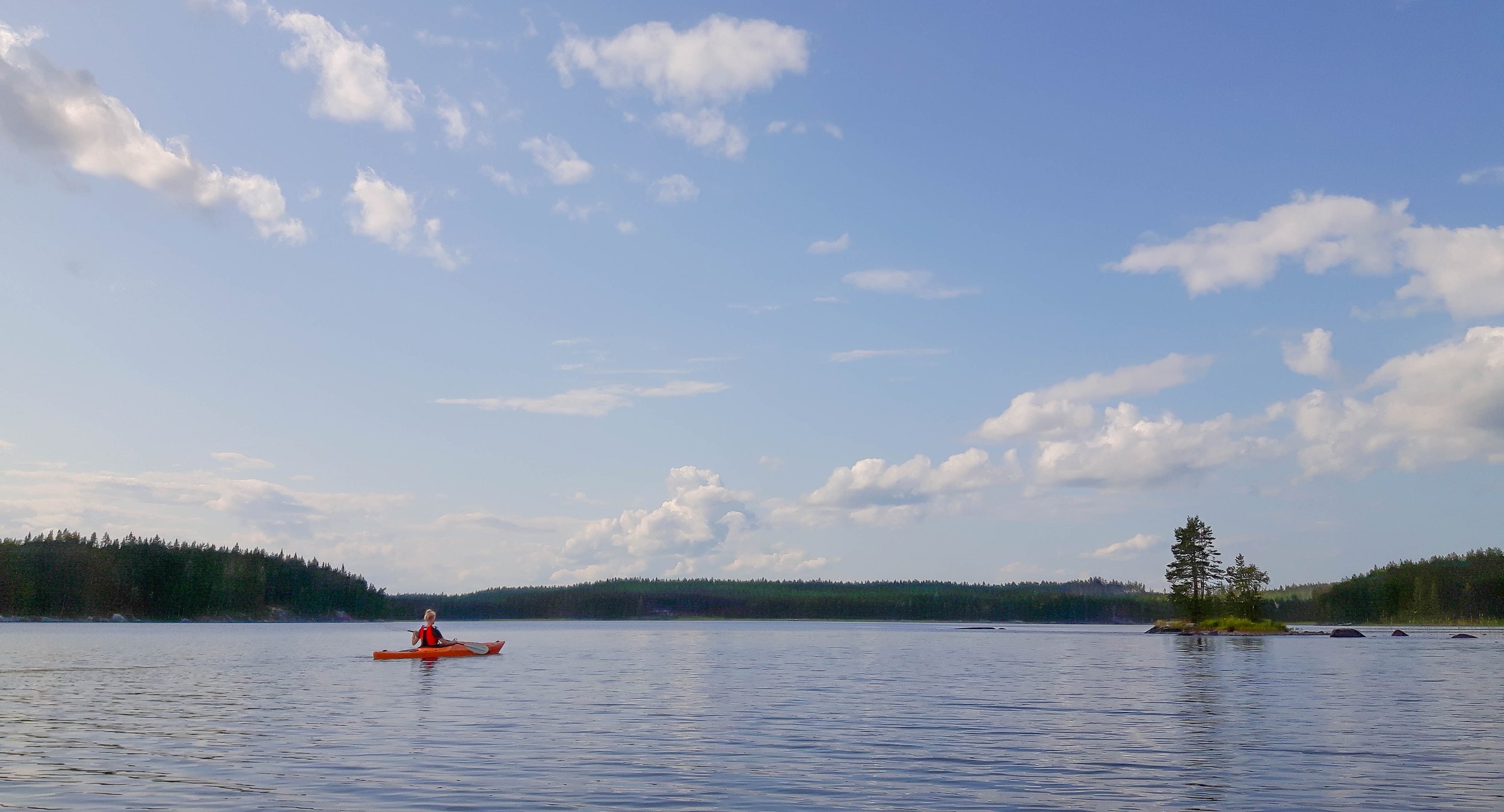 Henkilö meloo kanootilla järvellä, taustalla metsäinen rannat ja sininen taivas pilvillä.