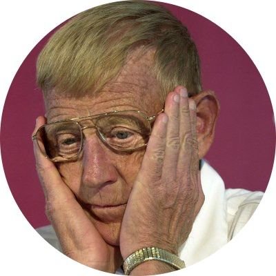 An elderly woman with short blonde hair and glasses, holding her head with both hands, appearing worried or distressed, with a pink background.