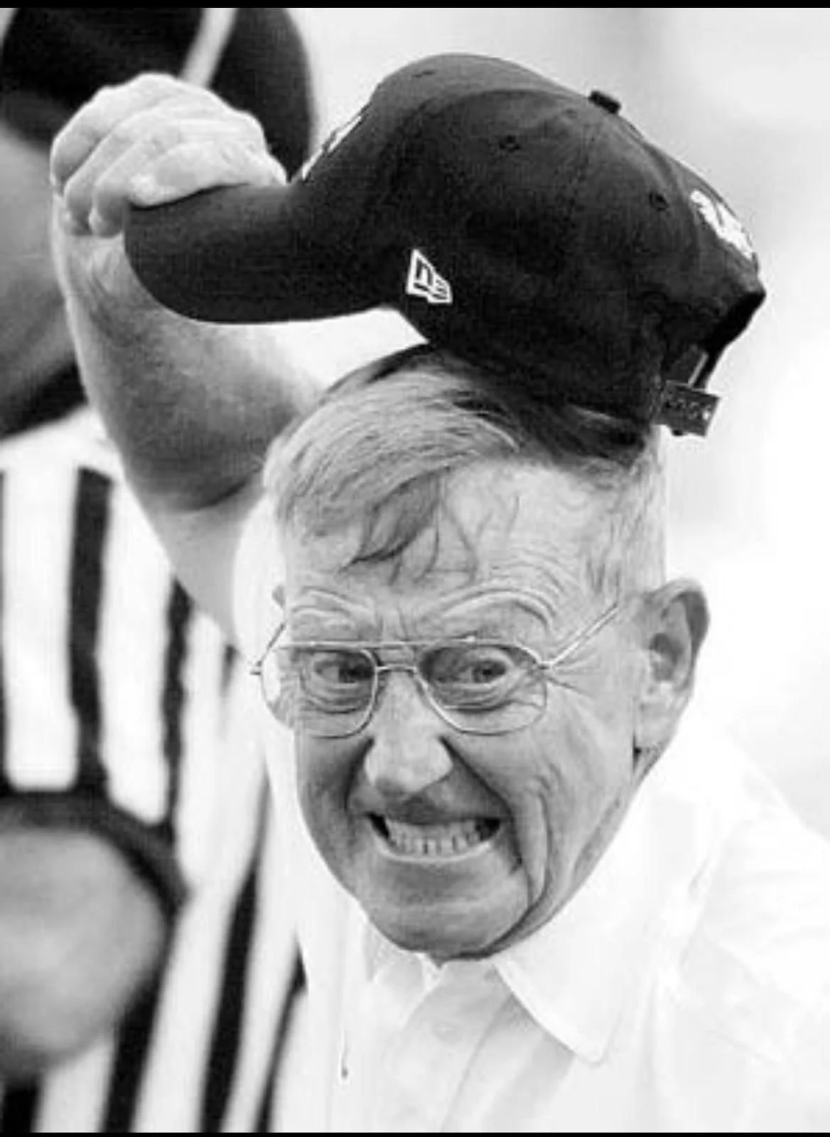 An elderly man with glasses grinning and looking at the camera, holding a black cap with a logo in his hand