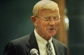 An elderly man with glasses and blonde hair speaking at a microphone, dressed in a dark suit and tie.