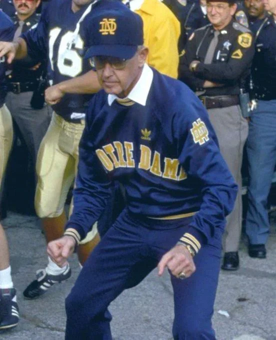 A man wearing sunglasses, a Notre Dame cap, and an Ole Miss sweatshirt dancing at a gathering with other people, including police officers.