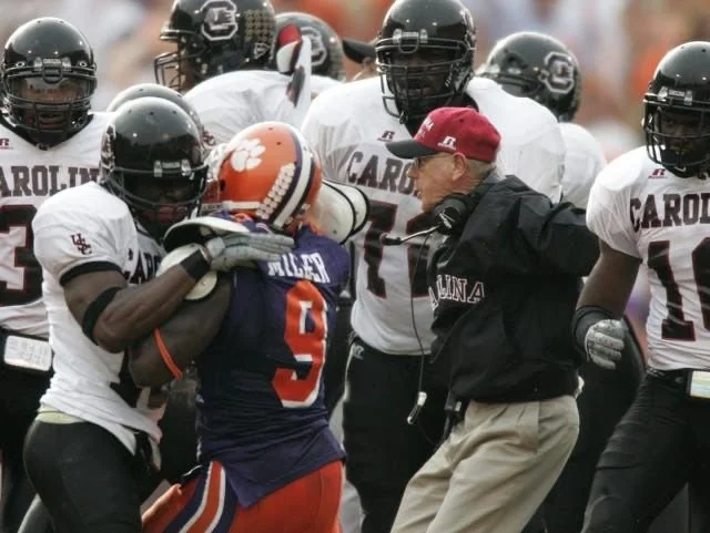 Football players from University of South Carolina and Clemson University, with a coach, during a game or practice, in an outdoor stadium.