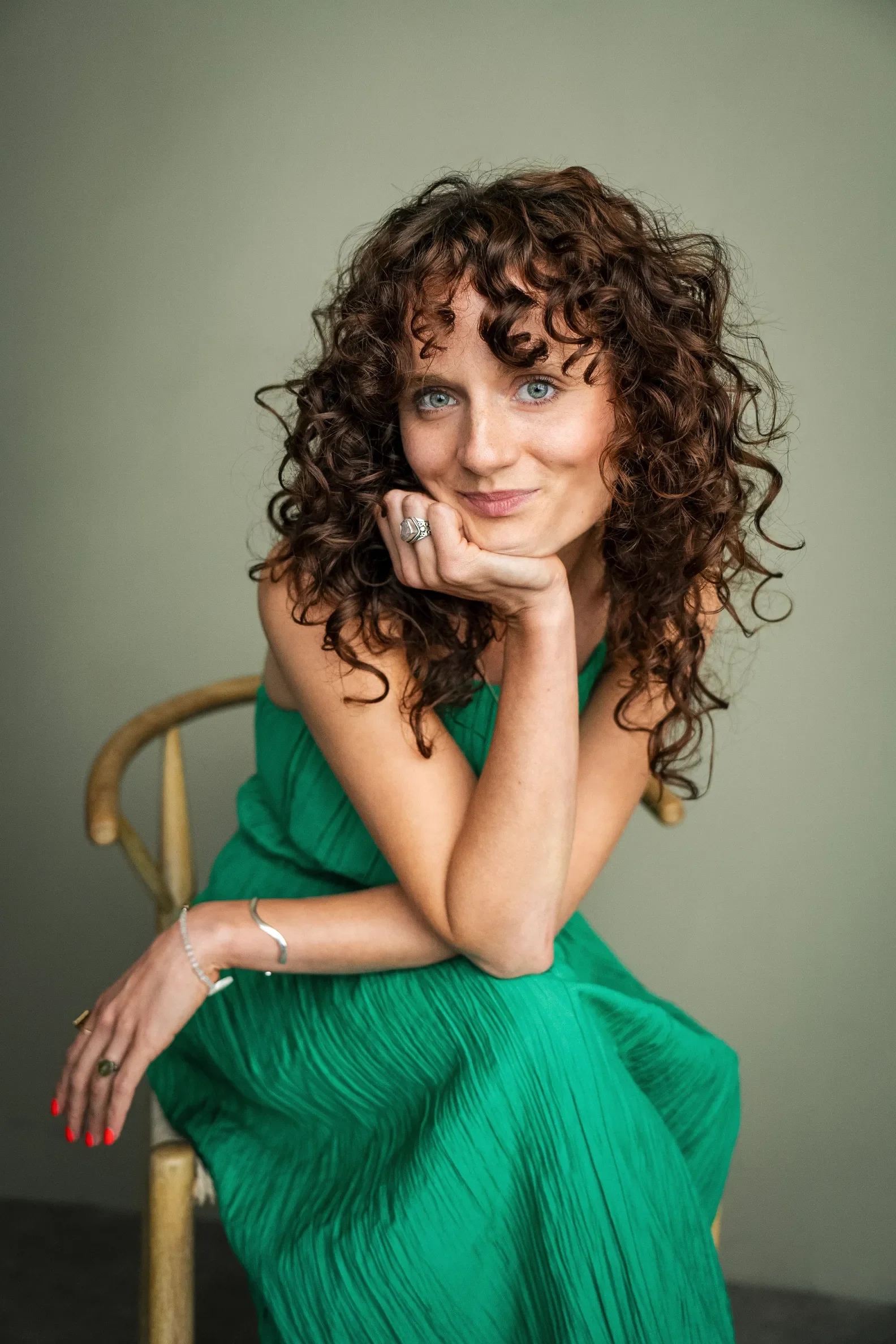A woman with curly brown hair wearing a green dress, seated on a wooden chair, resting her chin on her hand, looking at the camera with a slight smile.