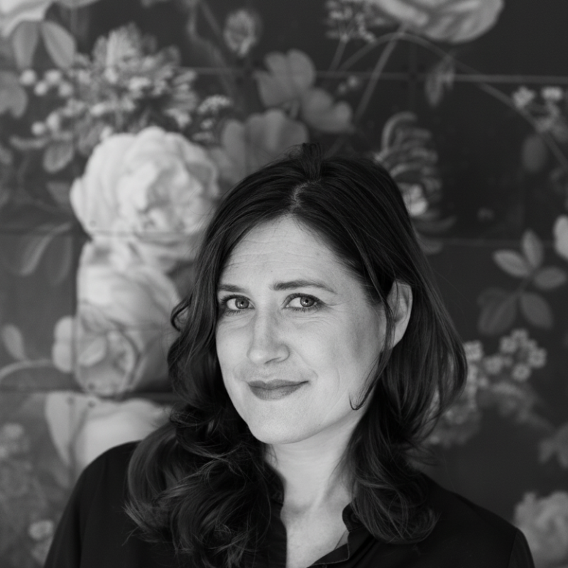 A woman with wavy dark hair smiling in front of a floral wallpaper background.