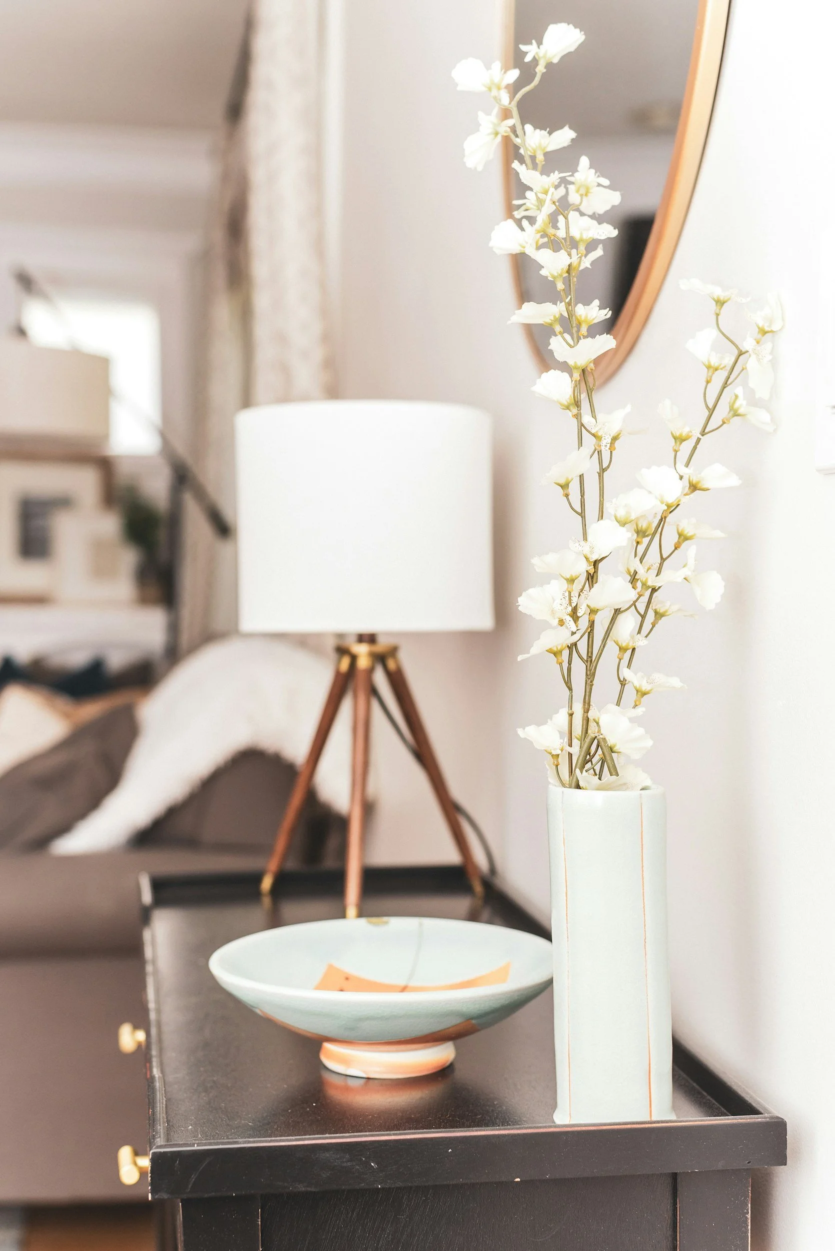 Vase blanc avec des branches de fleurs blanches sur une table noire, lampe avec pied en bois et abat-jour blanc, miroir rond en arrière-plan dans un intérieur moderne.