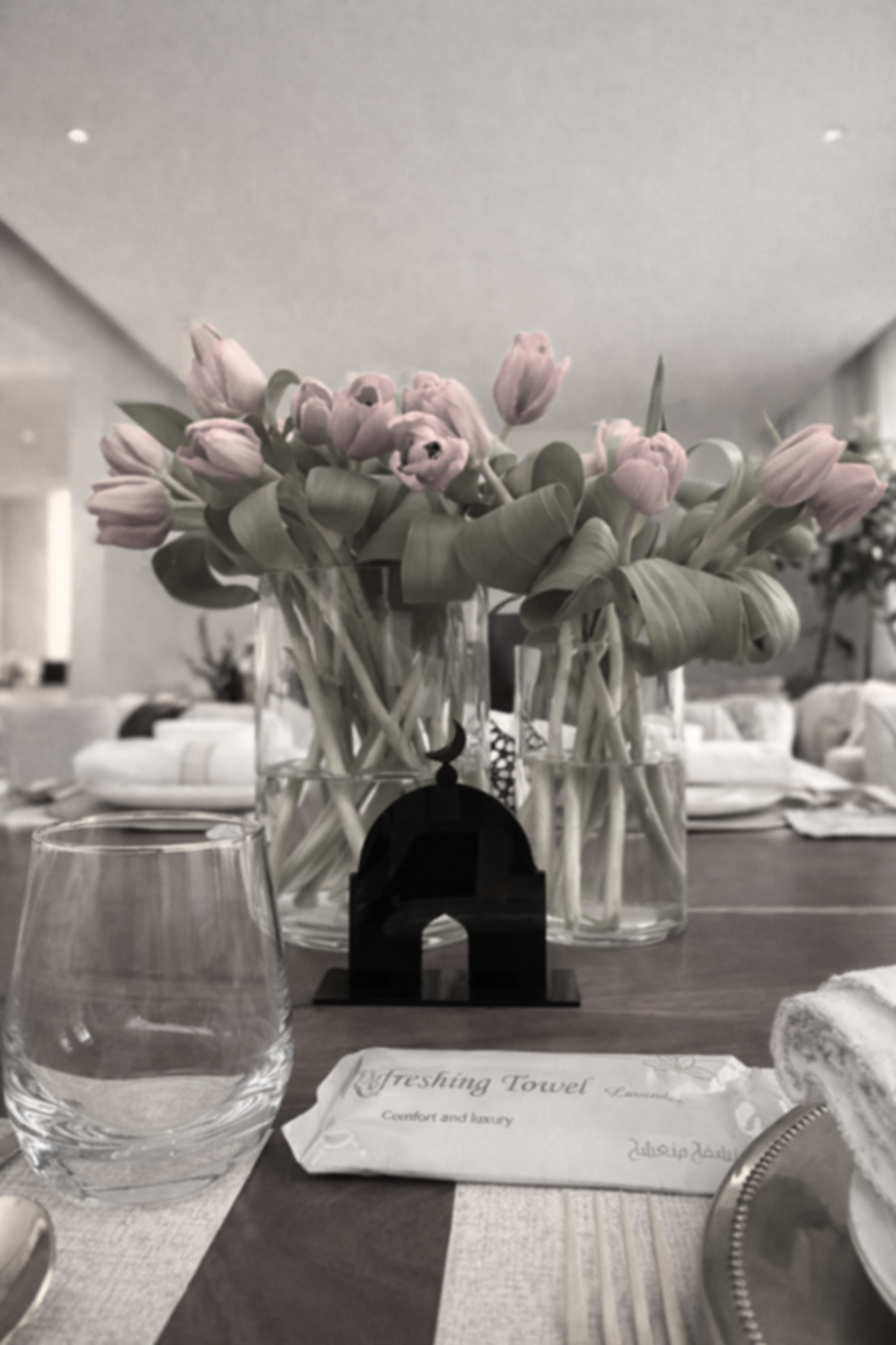 Un bouquet de tulipes roses dans un vase en verre sur une table a la maison, avec des verres d'eau, une serviette, un plateau, et un cadre noir au centre.