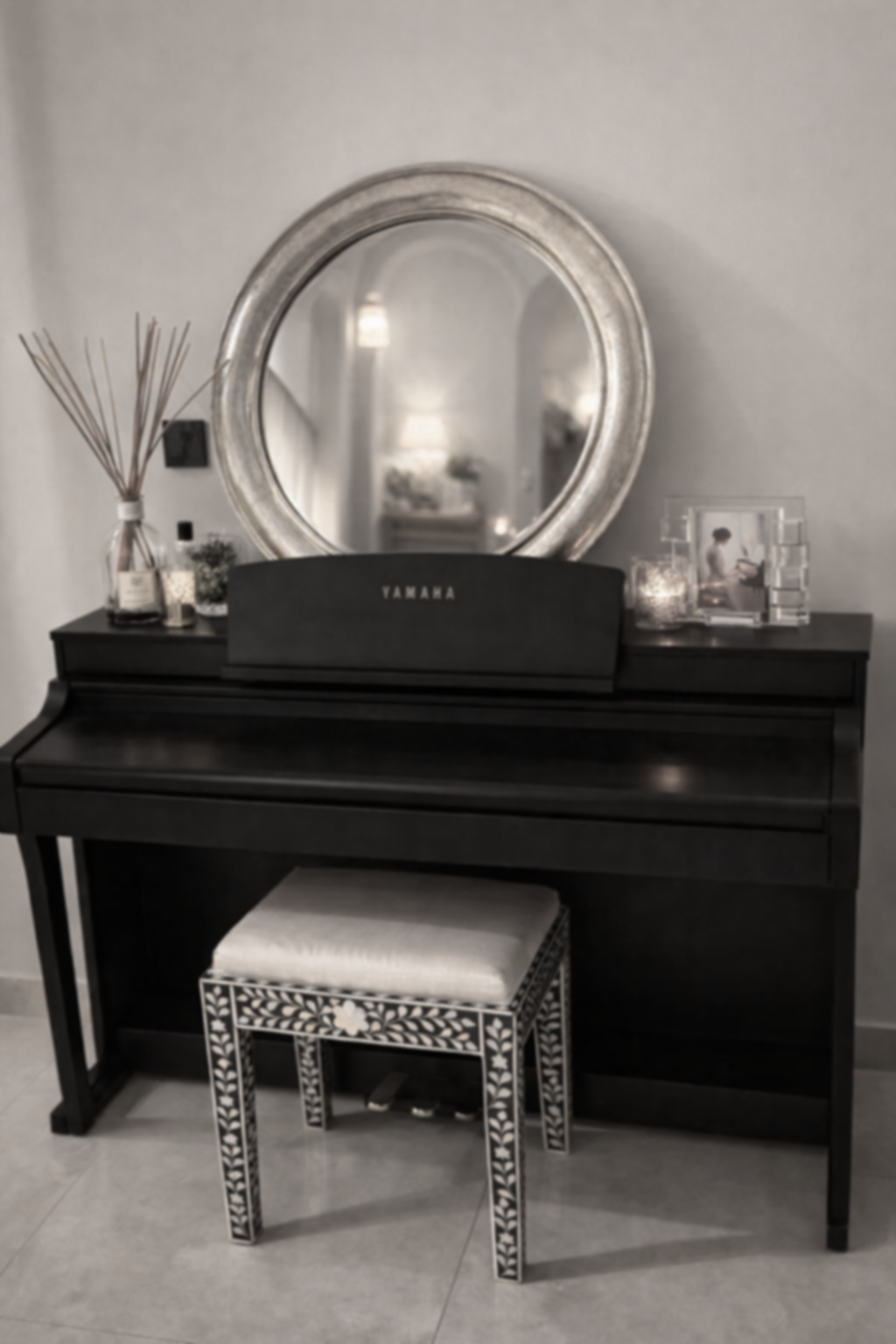Un piano noir avec un grand miroir rond argenté au mur et un tabouret décoratif devant. Sur le piano, il y a une bouteille avec des brindilles, un petit pot, une photo encadrée et deux bougies.