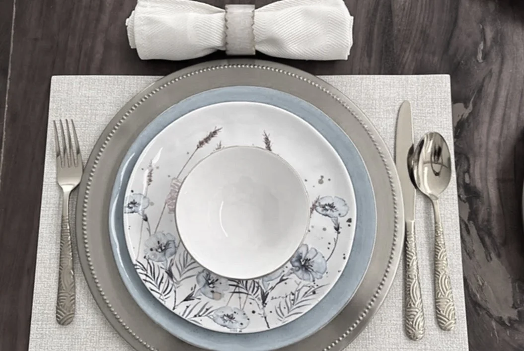 Une table dressée avec des assiettes décoratives, un couvert à gauche, un couteau et une cuillère à droite, une serviette enroulée en haut.