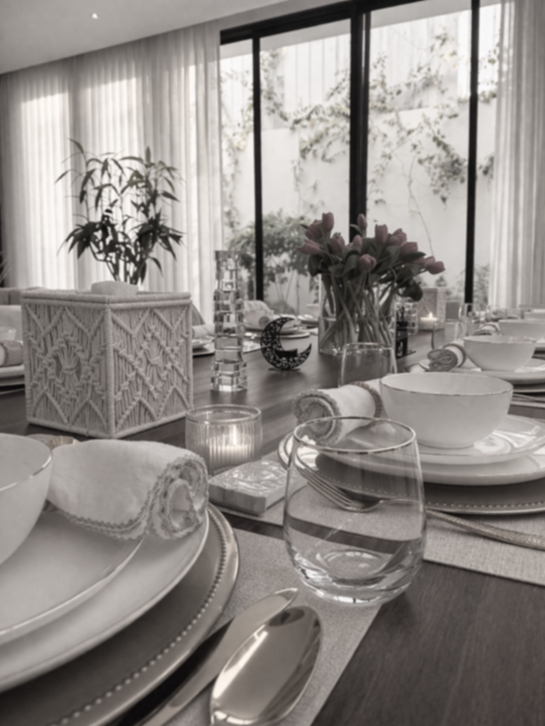 Une table de dîner élégante avec vaisselle blanche, verres en verre, serviettes roulées, et une vase avec des fleurs, dans une pièce lumineuse avec de grandes fenêtres et des rideaux.