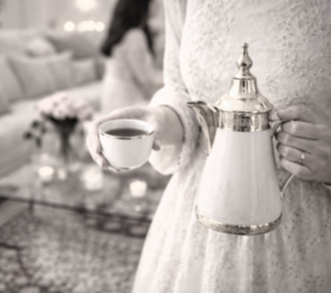 Une personne tient une tasse de café ou de thé dans une main et un service à thé dans l'autre, dans un intérieur décoré, avec un bouquet de fleurs en arrière-plan. 