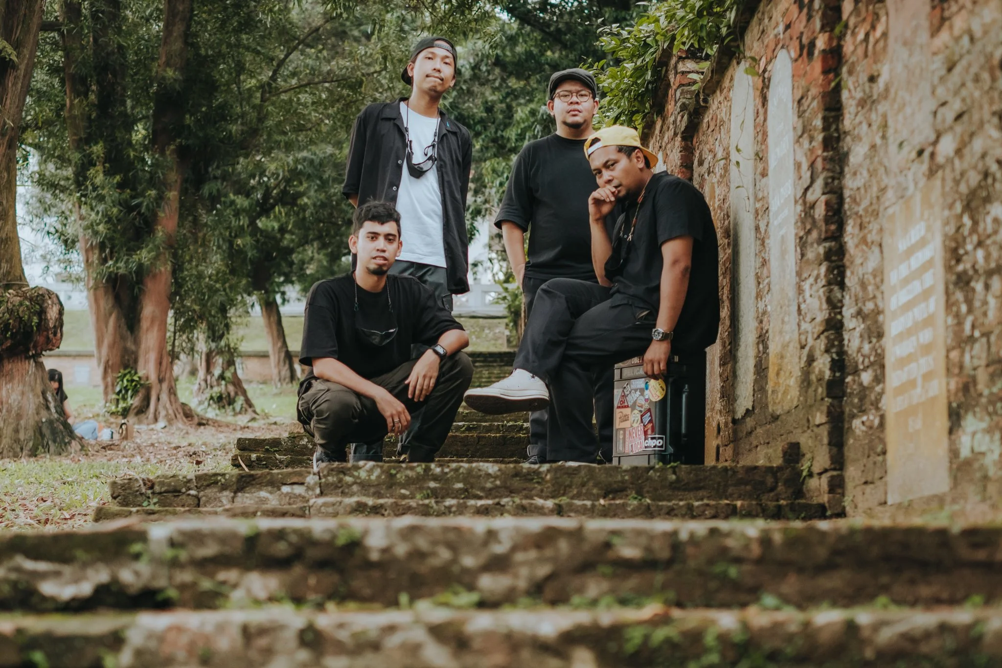 Five young men posing outdoors on stone steps near an old brick wall, with trees in the background.