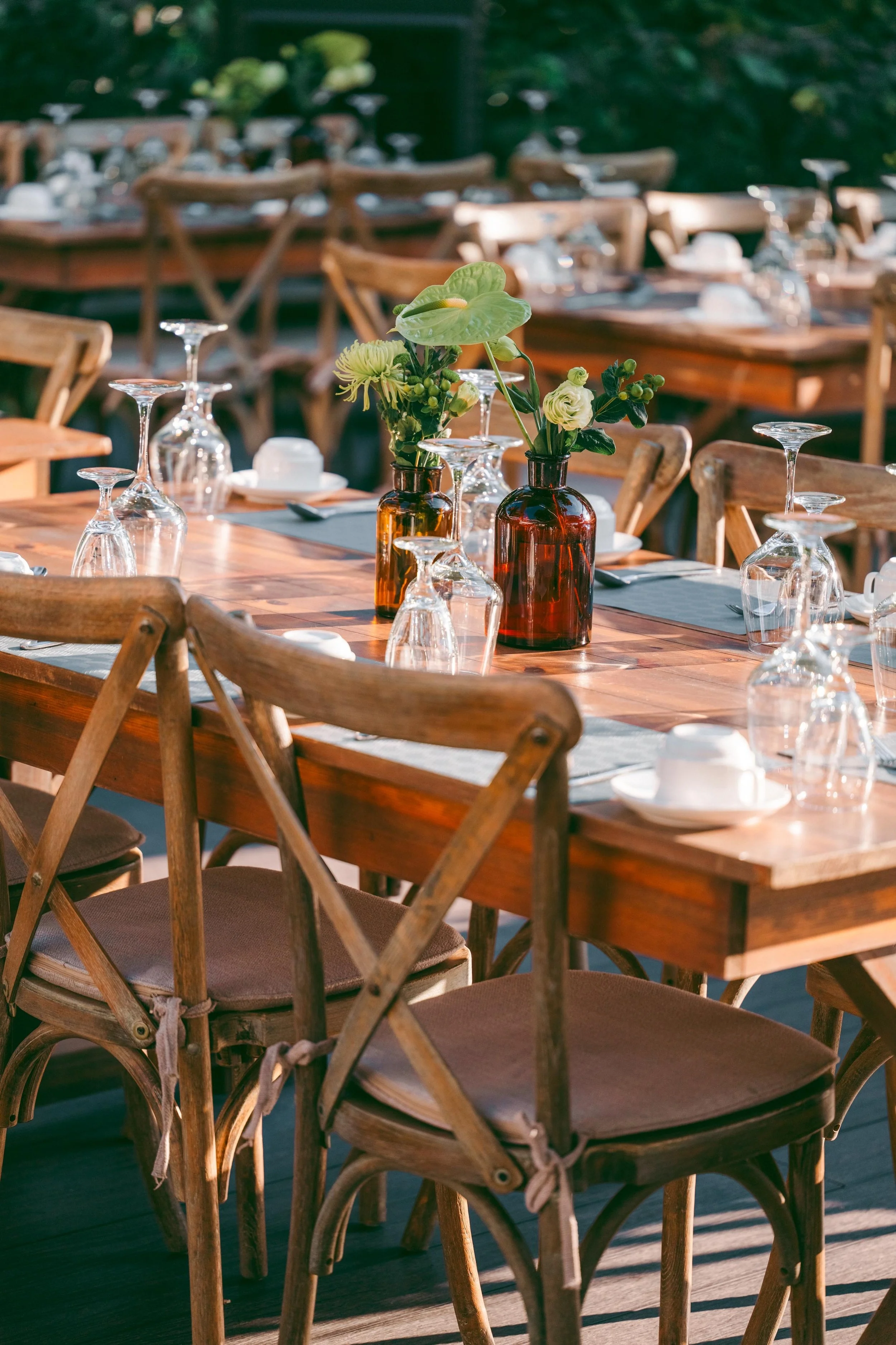 Elegant outdoor dining table set for a meal with glassware, plates, and floral centerpieces, surrounded by wooden chairs in sunlight.