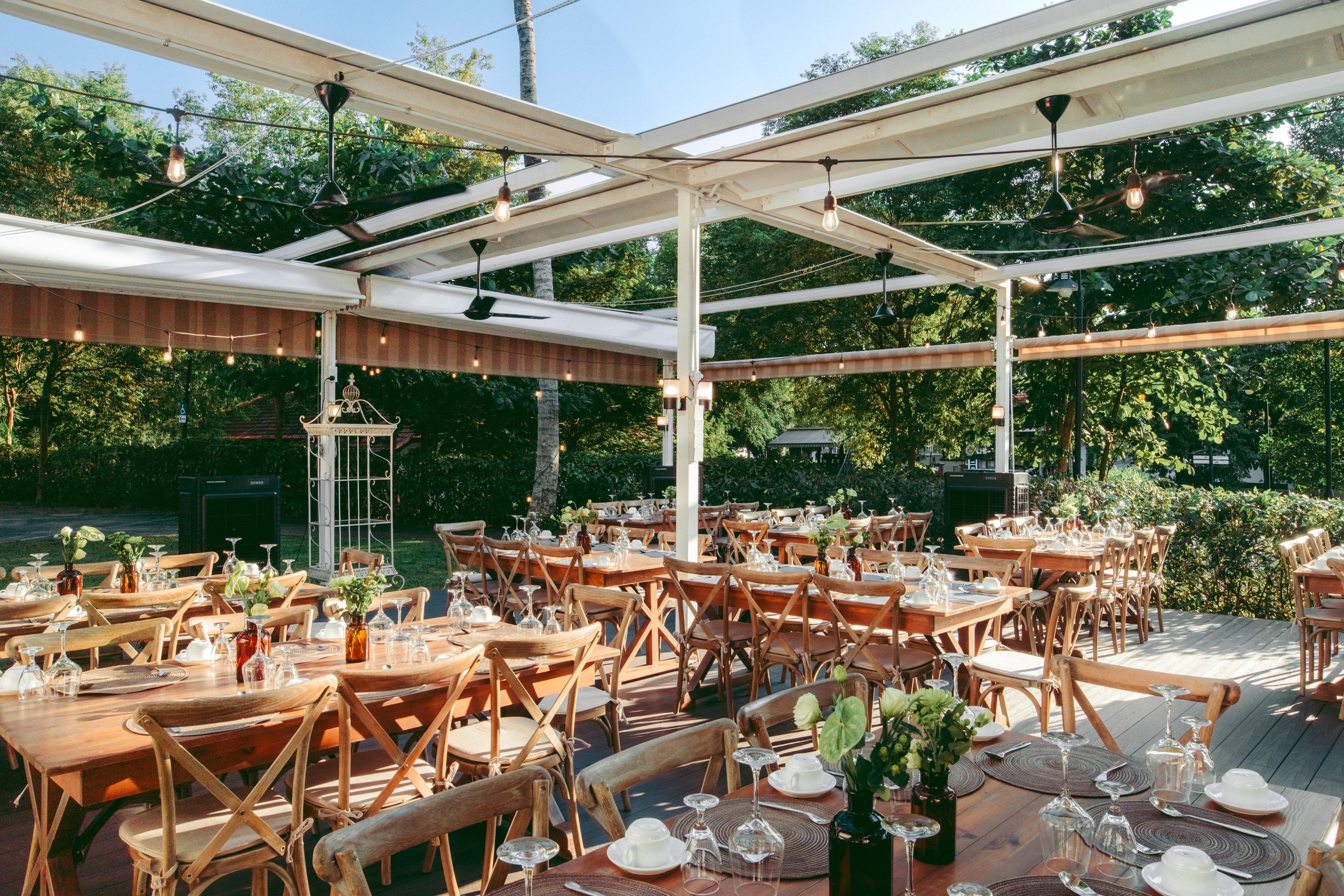 Outdoor dining area with wooden tables set with glassware, plates, and floral centerpieces, surrounded by wooden chairs, under a canopy with string lights, trees, and greenery in the background.