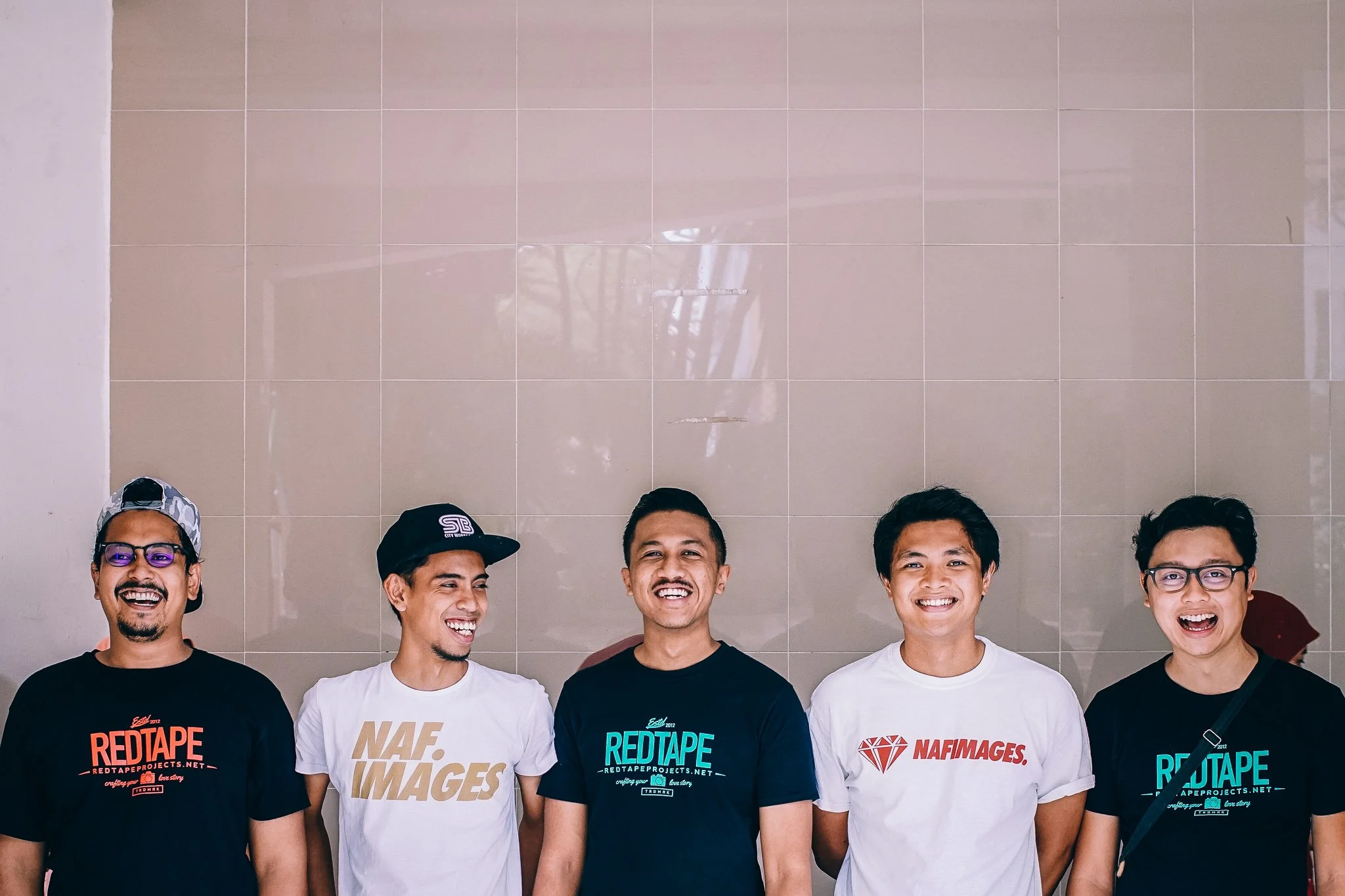 Five young men standing in front of a beige tiled wall, smiling and looking happy.