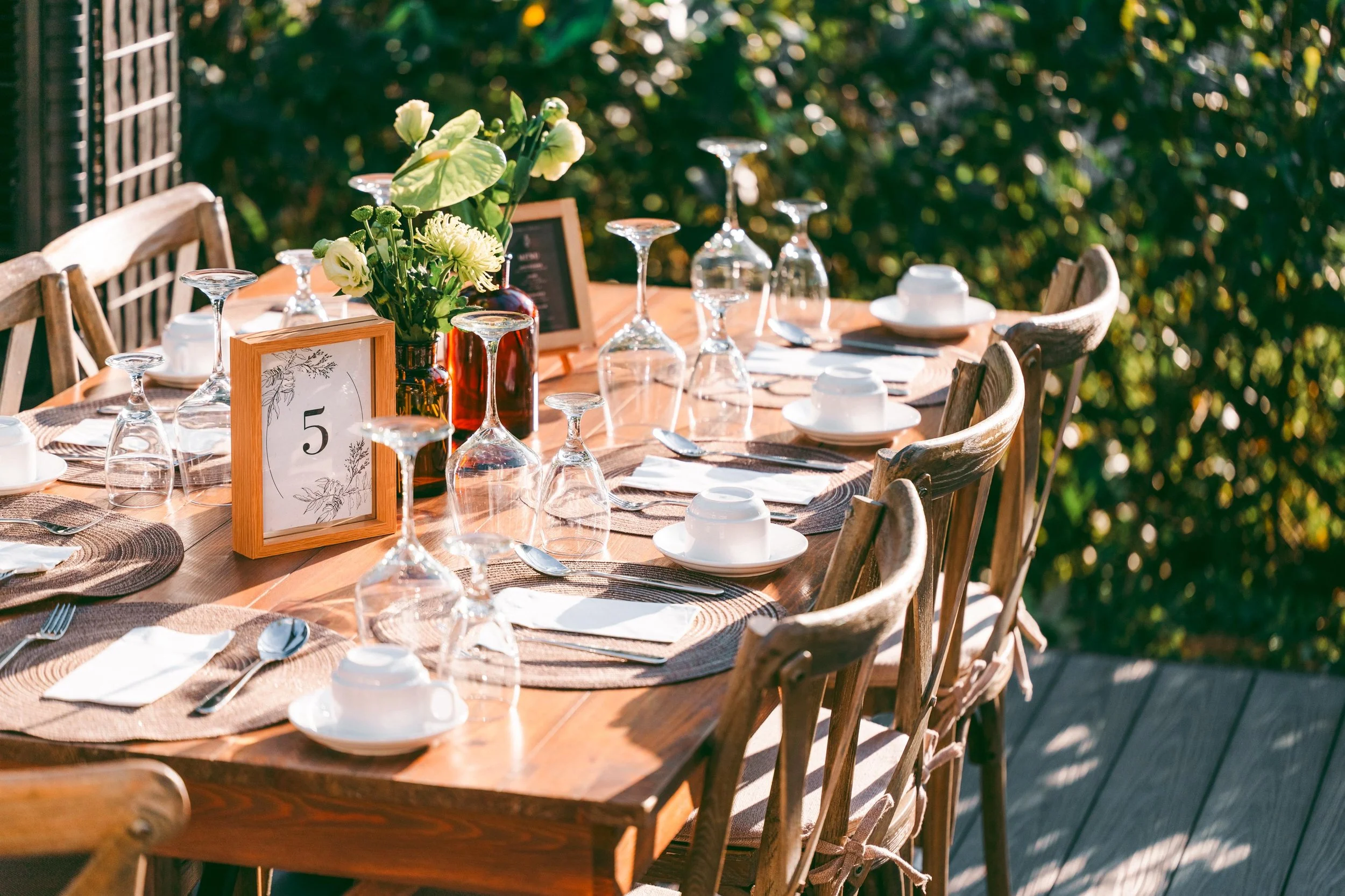 Outdoor dining table set with glasses, white bowls, silverware, a floral centerpiece, table number 5, and chairs, surrounded by greenery and sunlight.