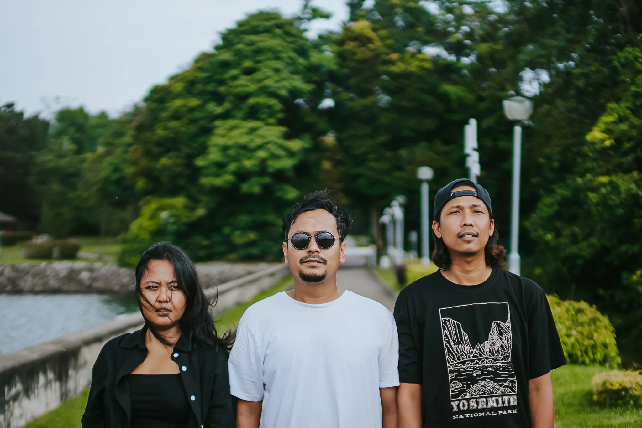 Three young adults standing outdoors near a river and large green trees, with a walkway and lampposts in the background.