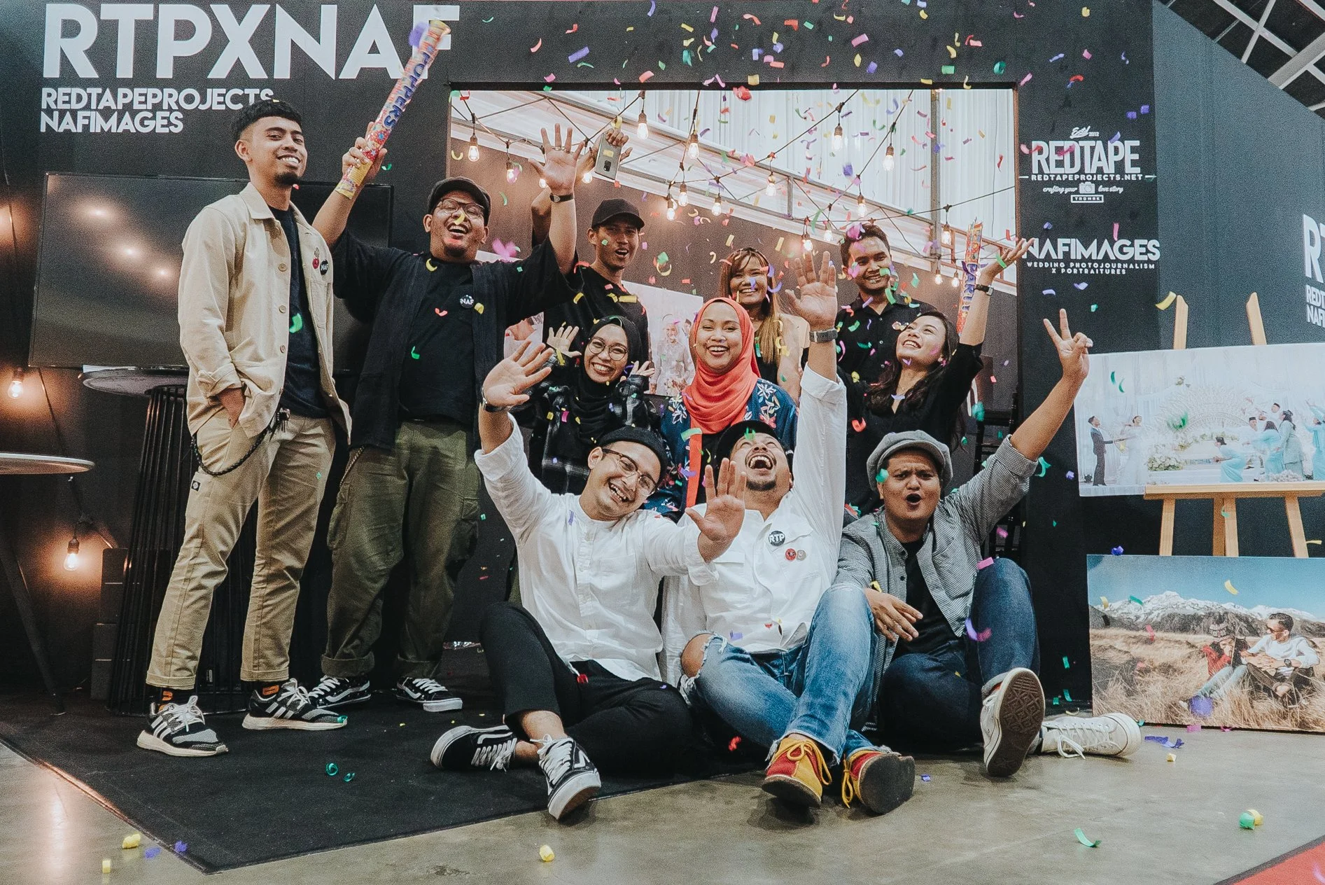 Group of people celebrating with confetti, smiling, some sitting on the floor and others standing on a stage at an event.