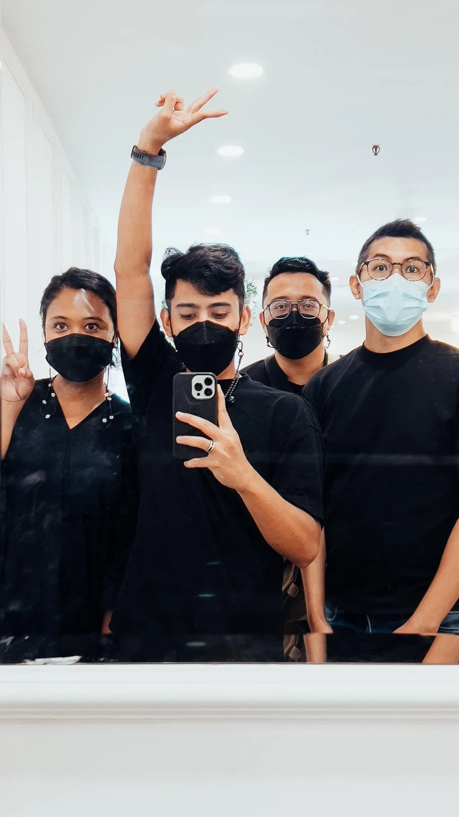 A group of four diverse young adults taking a selfie in front of a mirror, all wearing black shirts and face masks in a well-lit indoor setting.