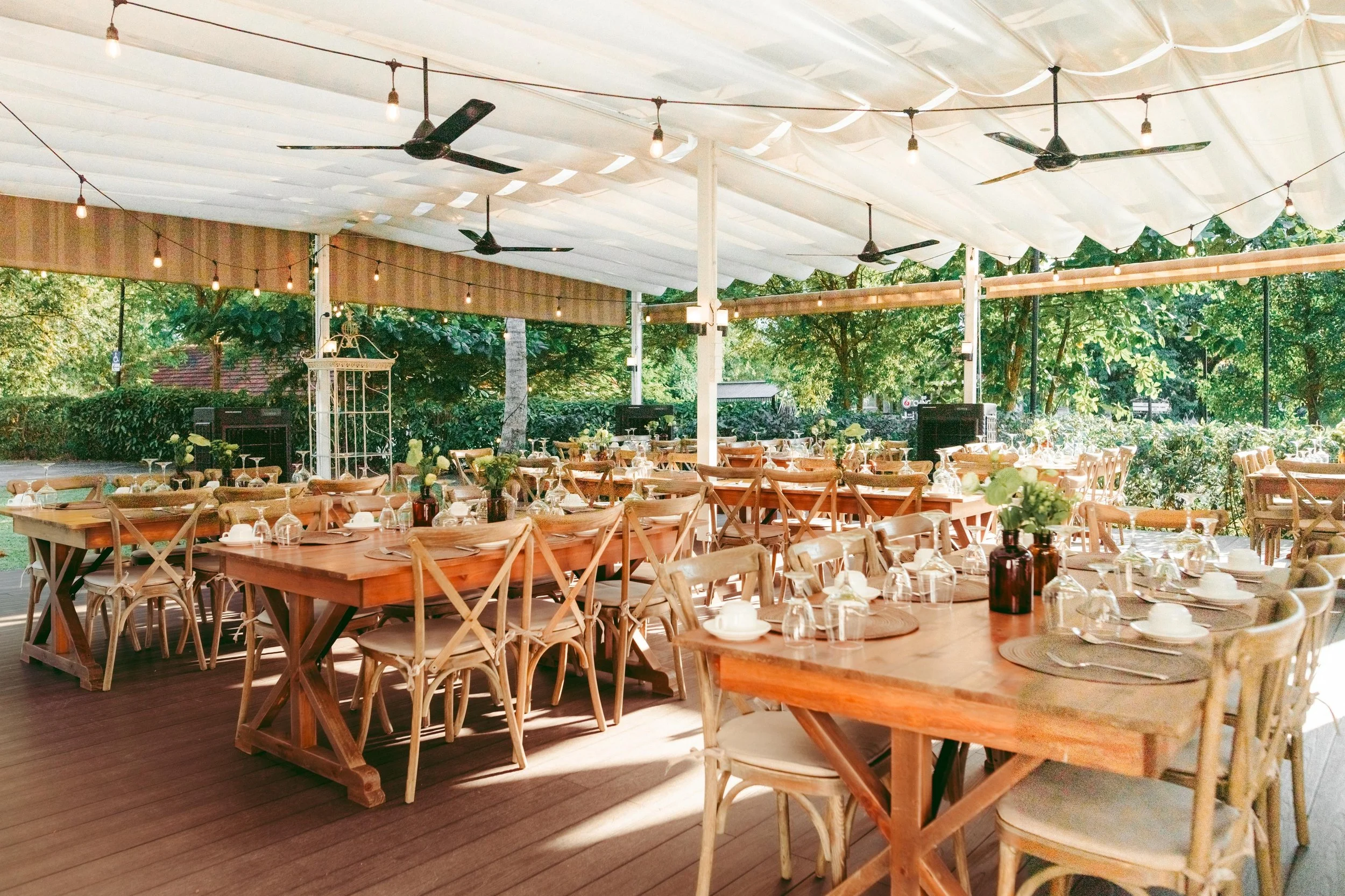 An outdoor dining area with wooden tables and chairs under a white tent with string lights, surrounded by greenery.