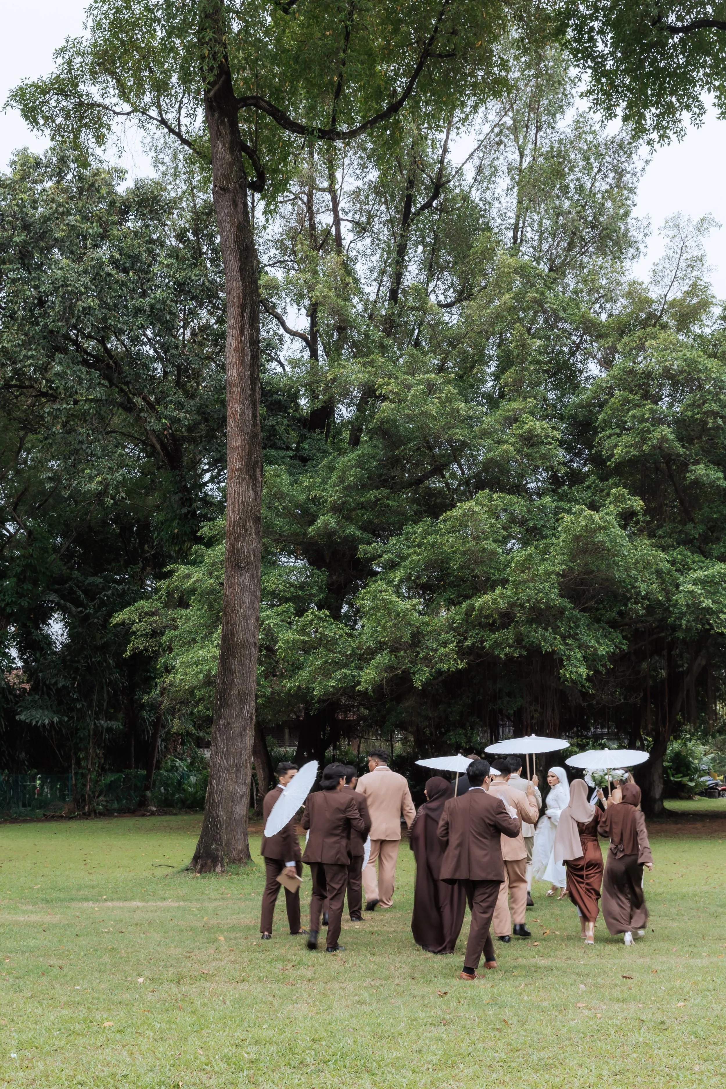 Group of people walking on a grassy lawn with umbrellas, surrounded by tall trees in a park or outdoor setting.