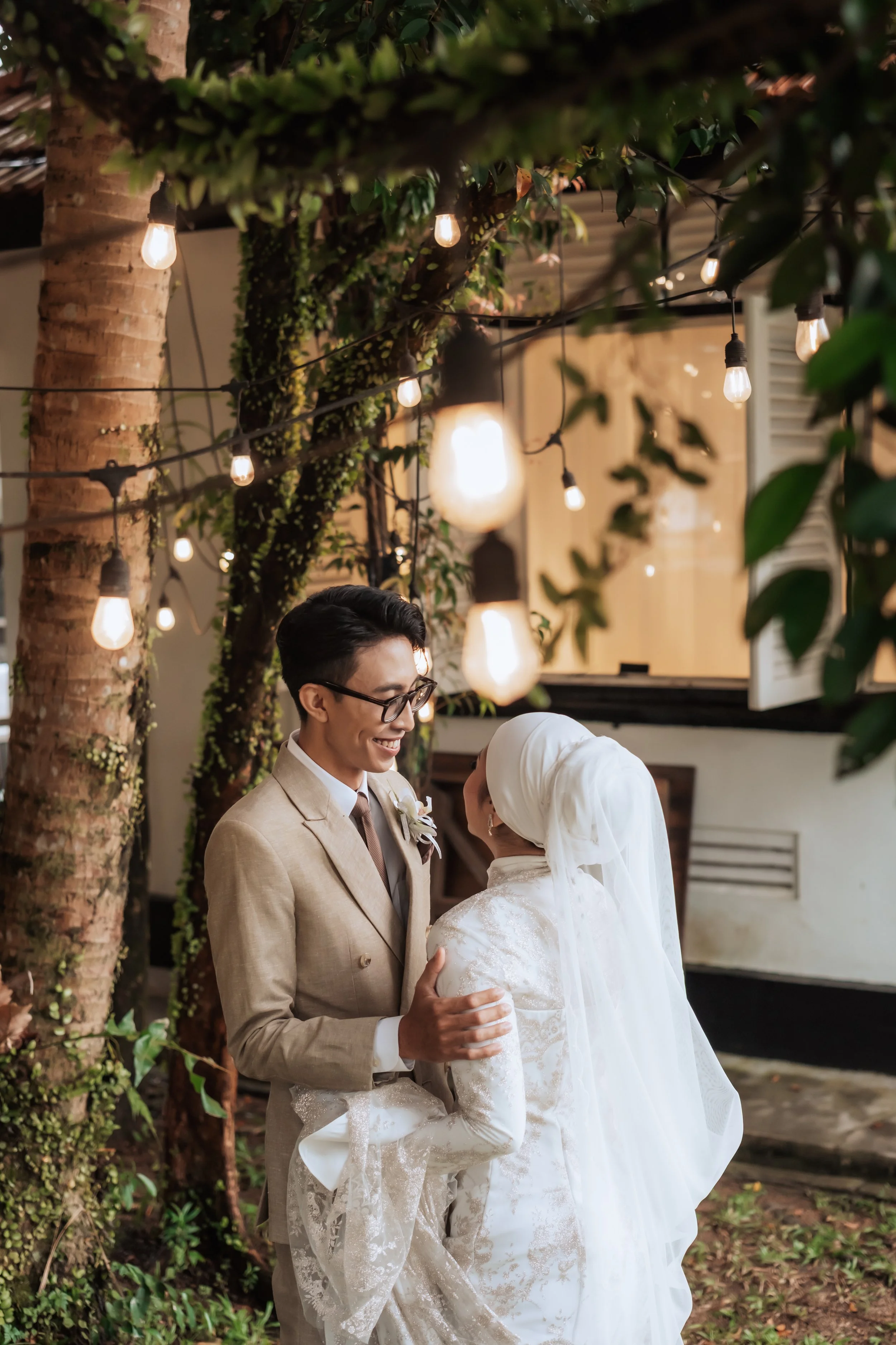 A happy bride and groom share a moment under string lights outdoors during their wedding reception. The groom is dressed in a beige suit and glasses, and the bride wears a white dress with a white headscarf. They are smiling and holding each other.