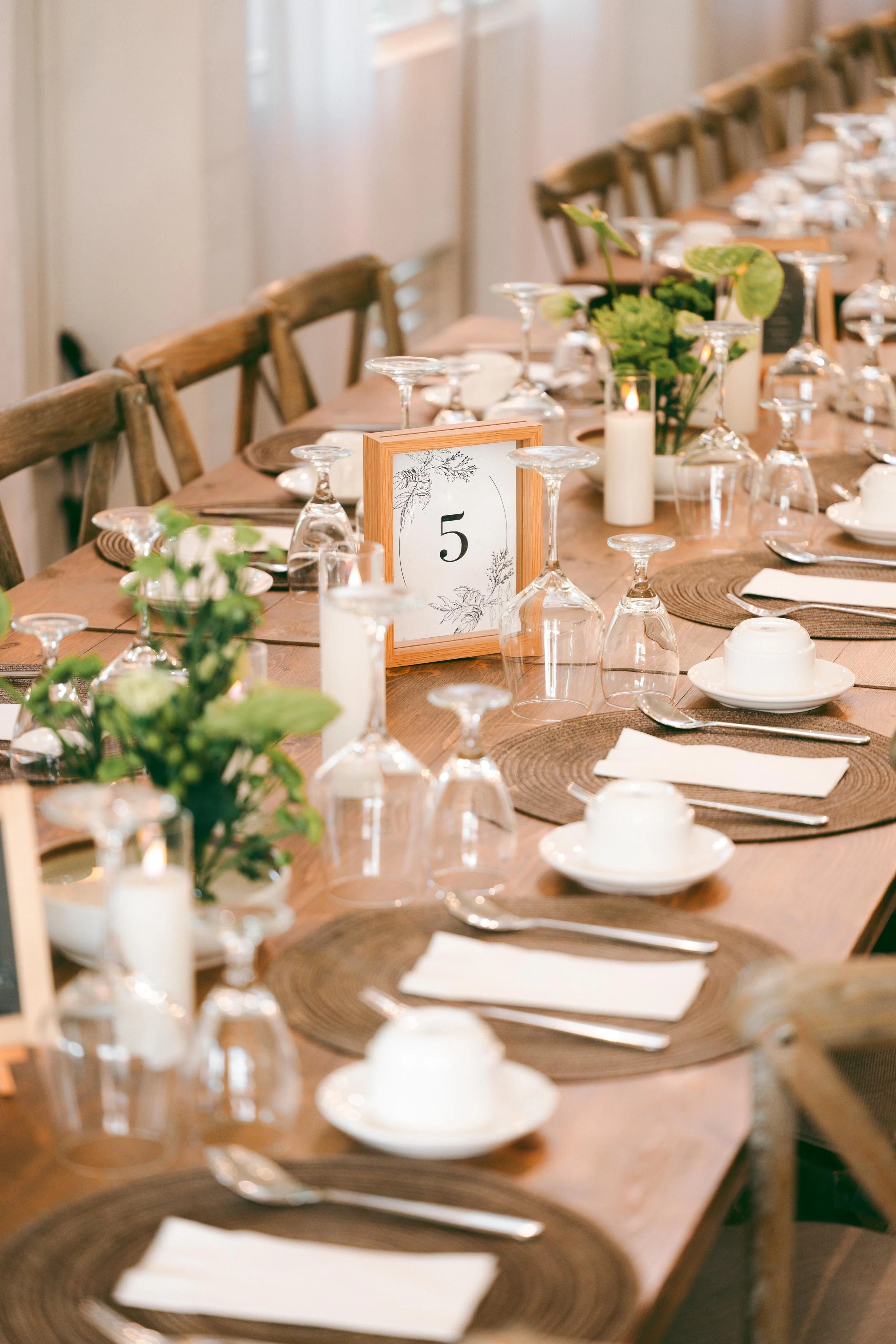 Elegant dinner table set with wine glasses, plates, silverware, candles, and greenery, with a table number five card in the center.