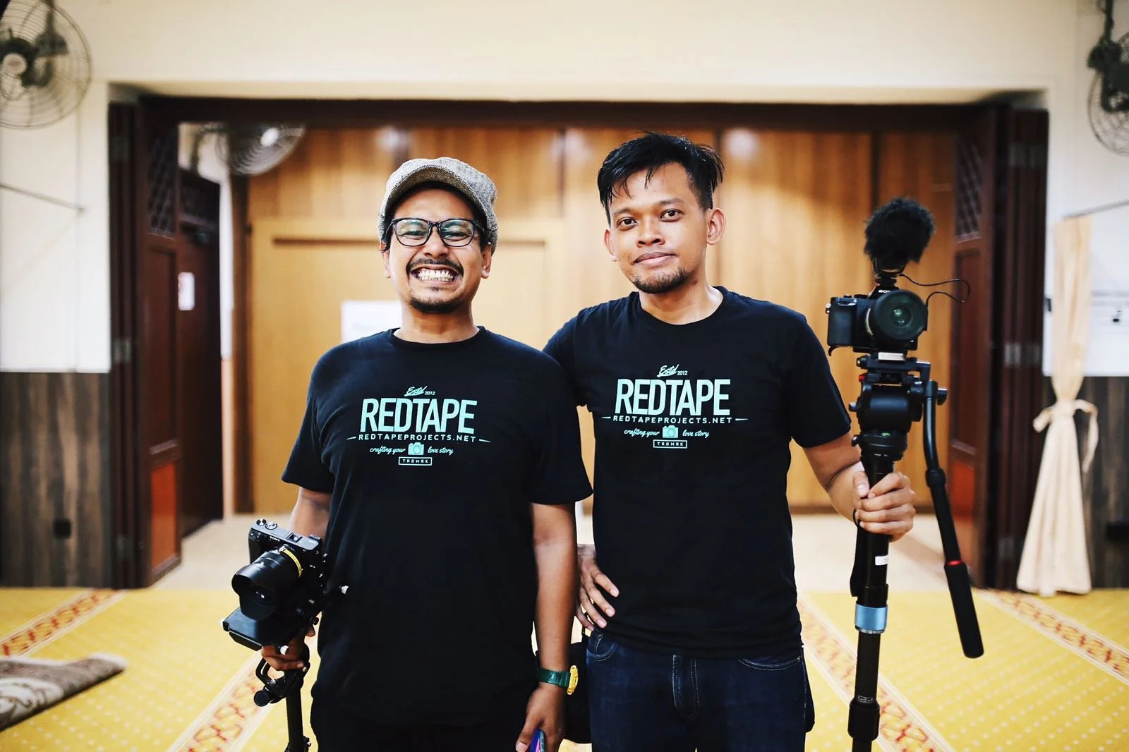 Two men wearing black T-shirts with 'Red Tape' logo, holding cameras, standing in a room with wooden walls, carpeted floor, and fans on the walls.