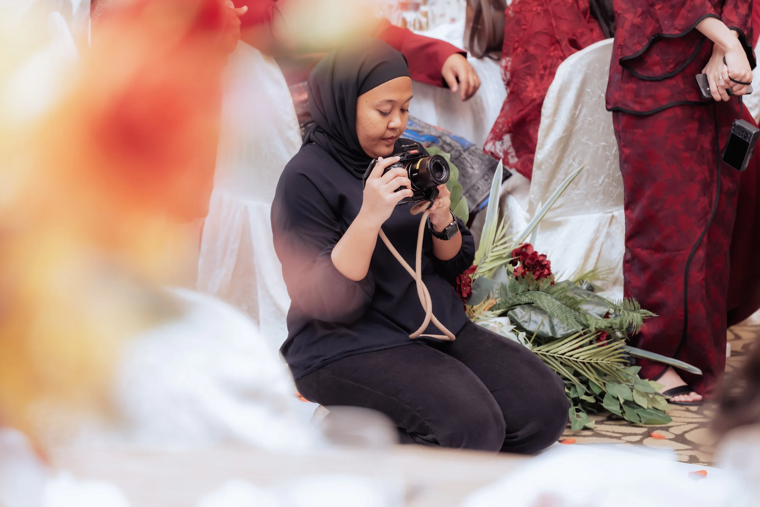 A woman wearing a black hijab and black clothing is sitting on the floor, focusing on her camera. She is surrounded by people in traditional attire and floral decorations, indicating a formal or cultural event.