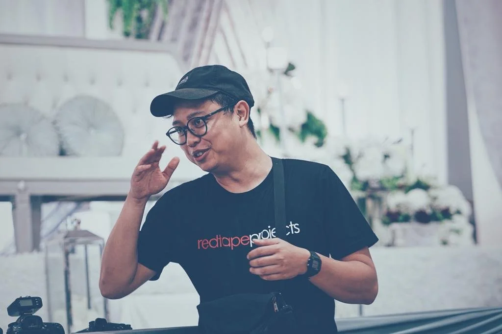 A man wearing glasses, a black cap, and a black t-shirt with red and white text, standing indoors with a decorated background, gesturing with one hand near his face.