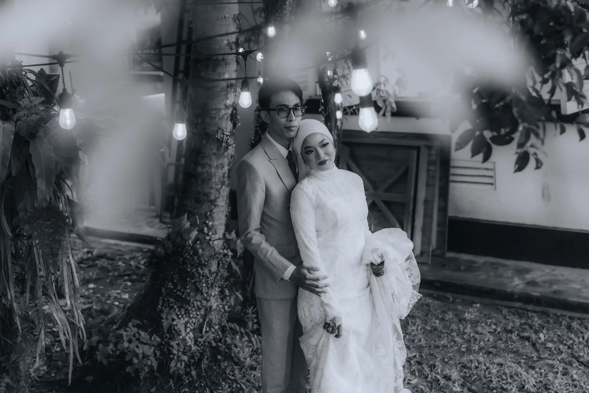 A black and white photo of a couple, the man in a light suit with glasses and the woman in a white wedding dress with a headscarf, standing outdoors near a tree with hanging light bulbs and foliage.