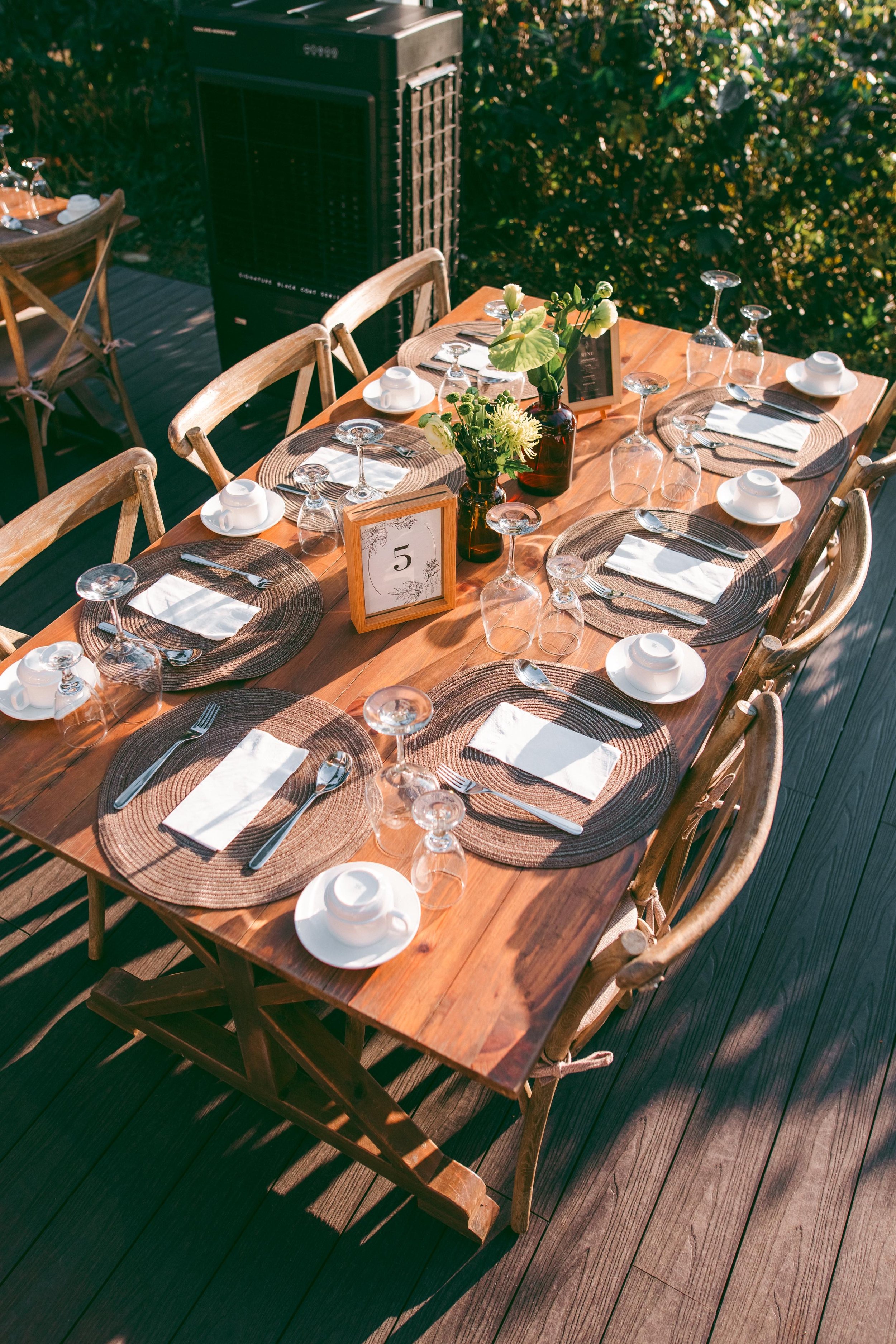 A neatly set outdoor dining table with six place settings, glassware, and napkins, decorated with a centerpiece of flowers and a table number, on a wooden deck surrounded by greenery.