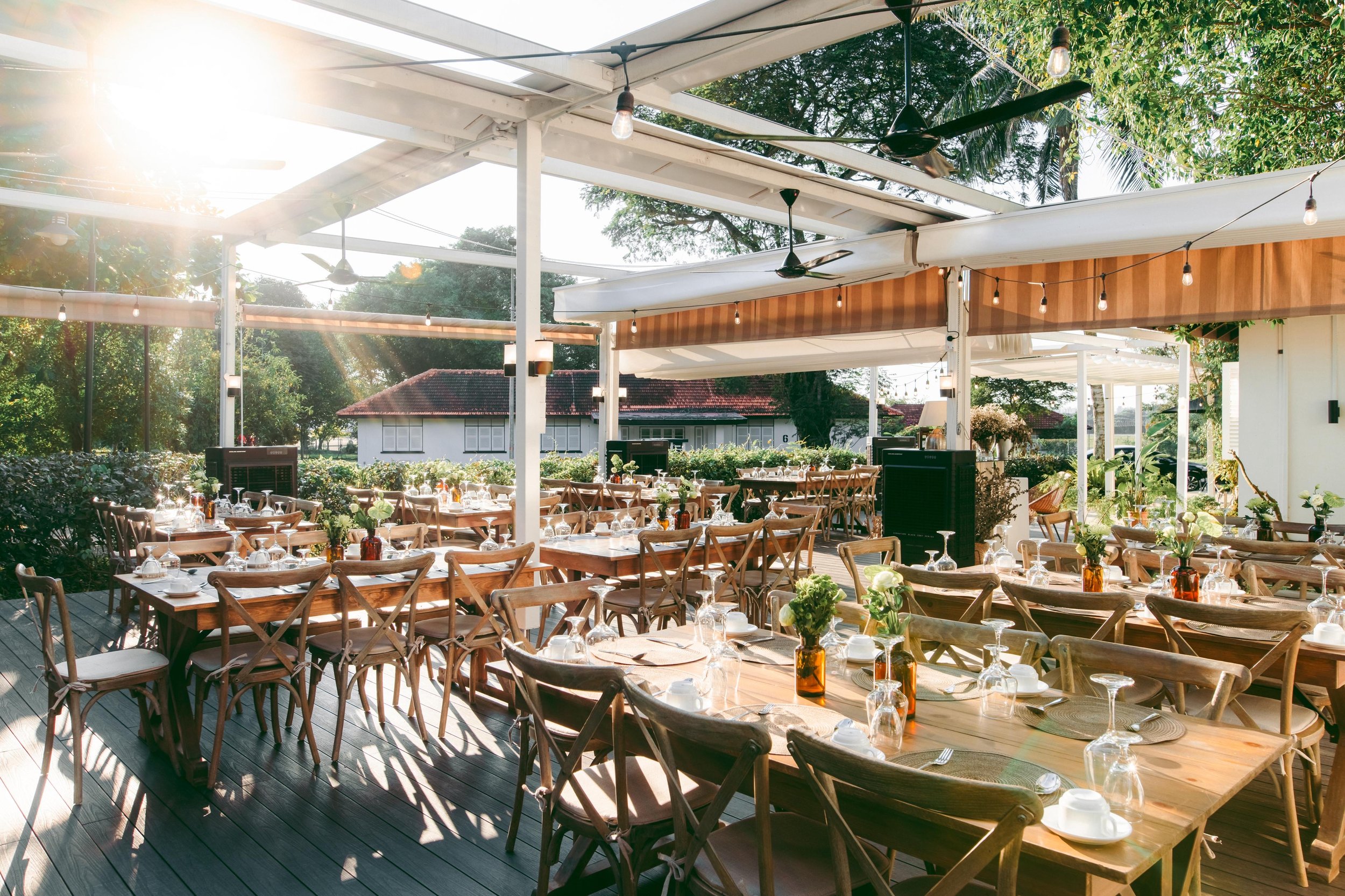 Outdoor restaurant patio with wooden tables set for dining, decorated with small flower vases, under a white canopy with string lights, ceiling fans, and surrounded by green trees and bushes.