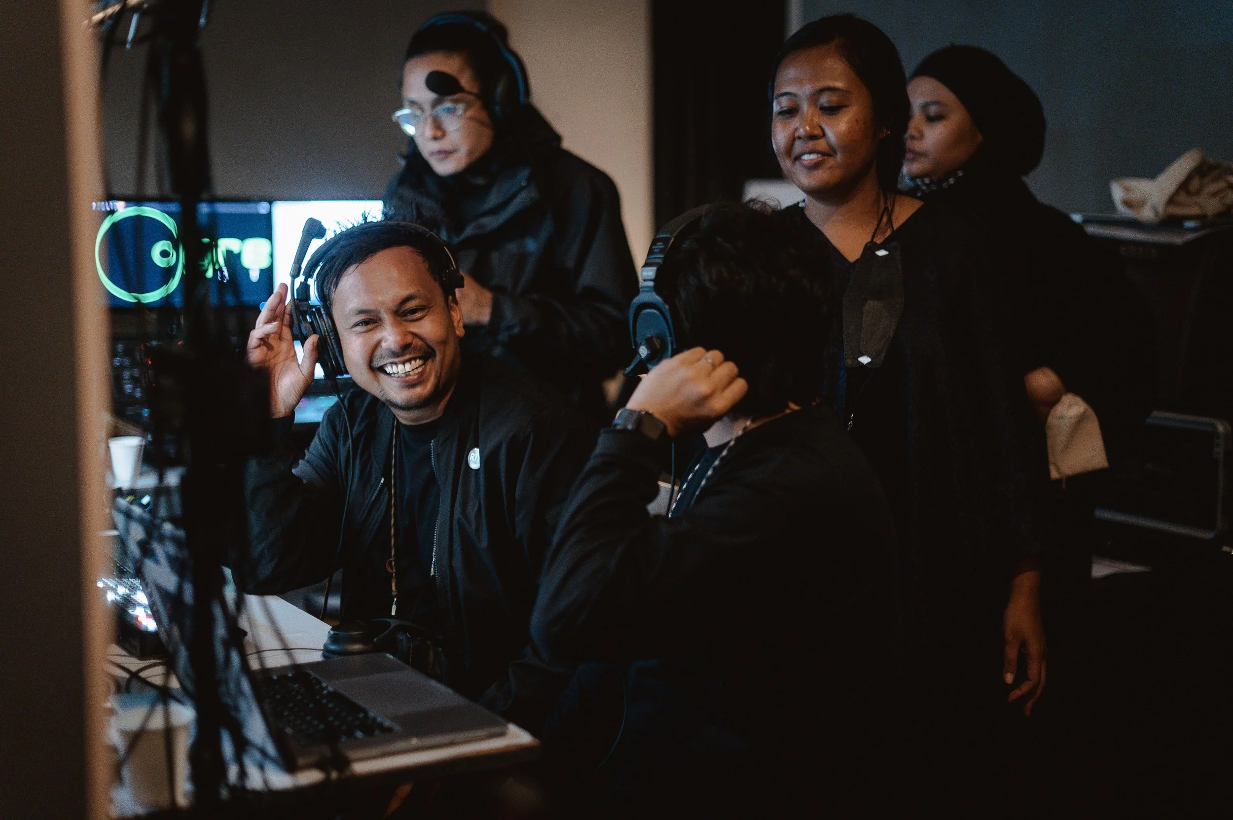 Group of diverse people in a dark room, some wearing headphones and smiling, working on computers, possibly at a gaming or tech event.