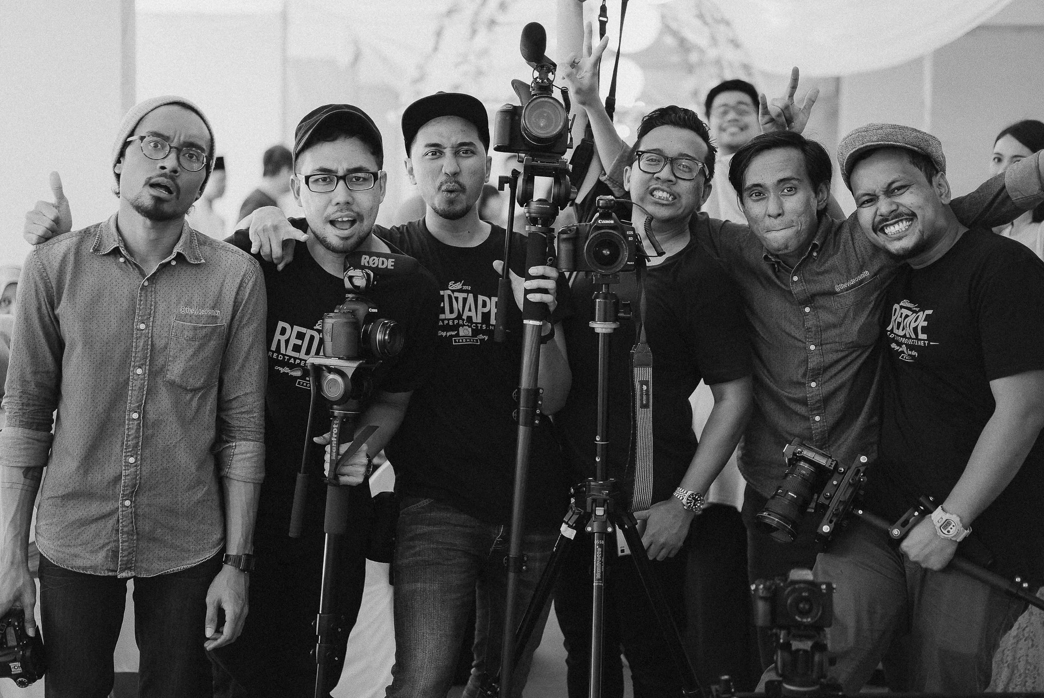 Group of seven men posing with cameras and equipment, some making expressive gestures, indoors with a large tent or canopy overhead.