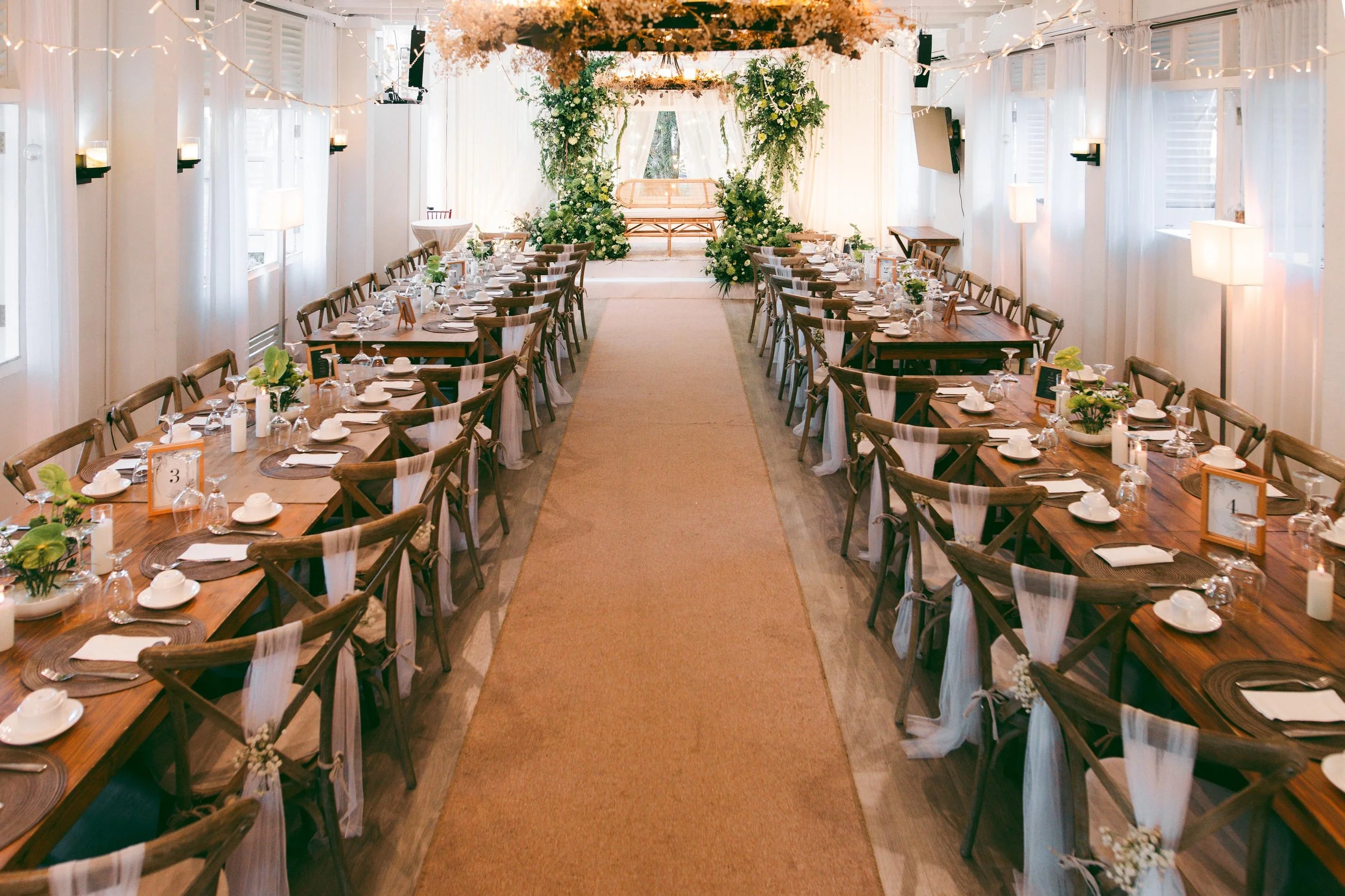 Elegant indoor wedding reception with long wooden tables on each side, decorated with white tableware and small floral centerpieces, set in a bright room with white curtains and string lights. A beige carpet runs down the middle leading to a floral a