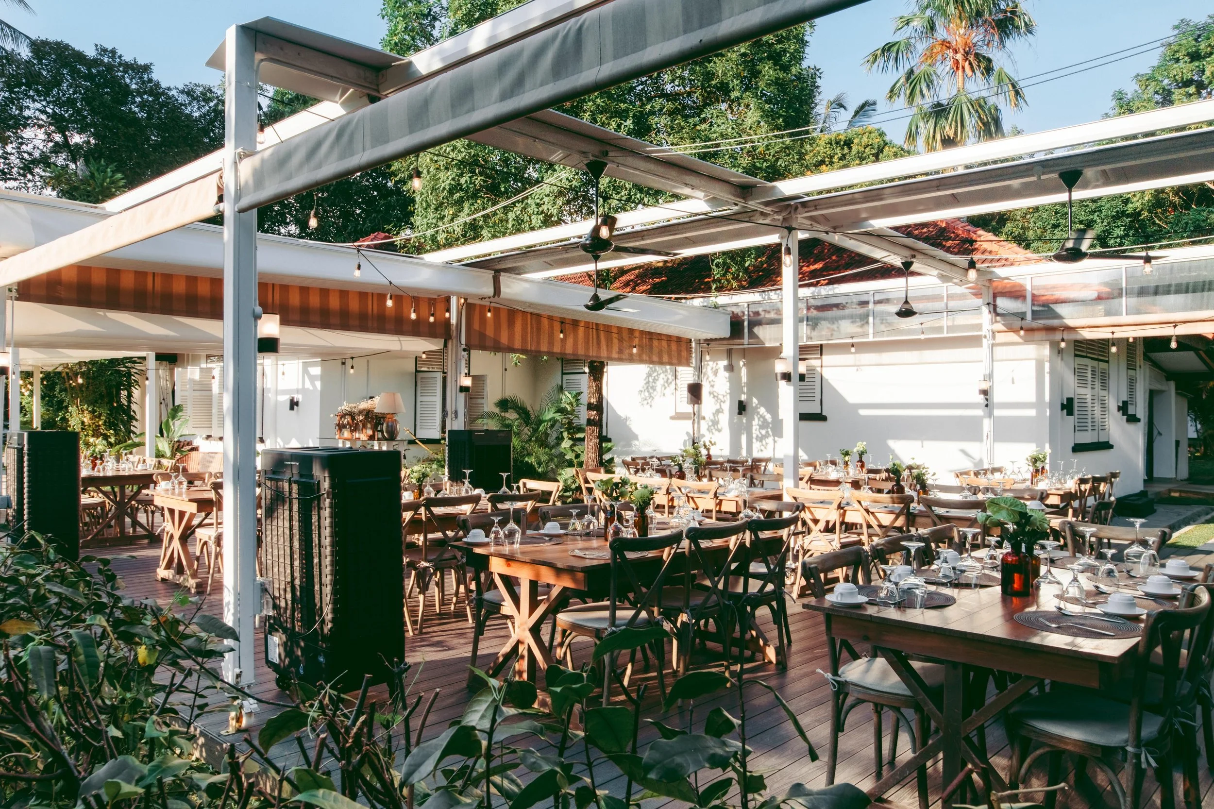 An outdoor restaurant or event space with wooden tables and chairs, set with plates and glasses, under string lights and a retractable awning, surrounded by greenery and palm trees, on a sunny day.