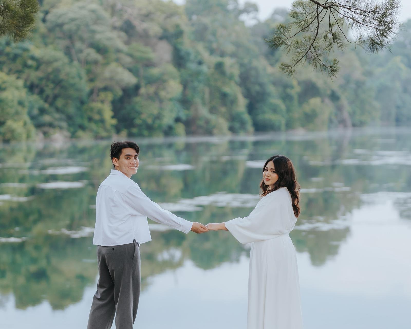A young man and woman holding hands and facing each other near a lake in a forested area, both dressed in white, with trees and water in the background.
