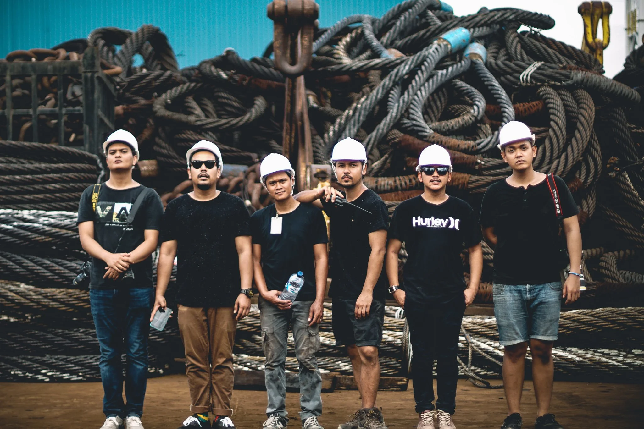 Six young men standing in front of a large pile of industrial ropes, wearing white safety helmets and casual clothing. One is holding a water bottle, and another has sunglasses.