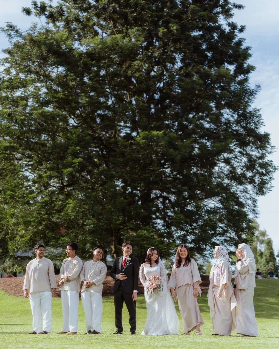 Couple in wedding attire standing with friends outdoors near a large tree.