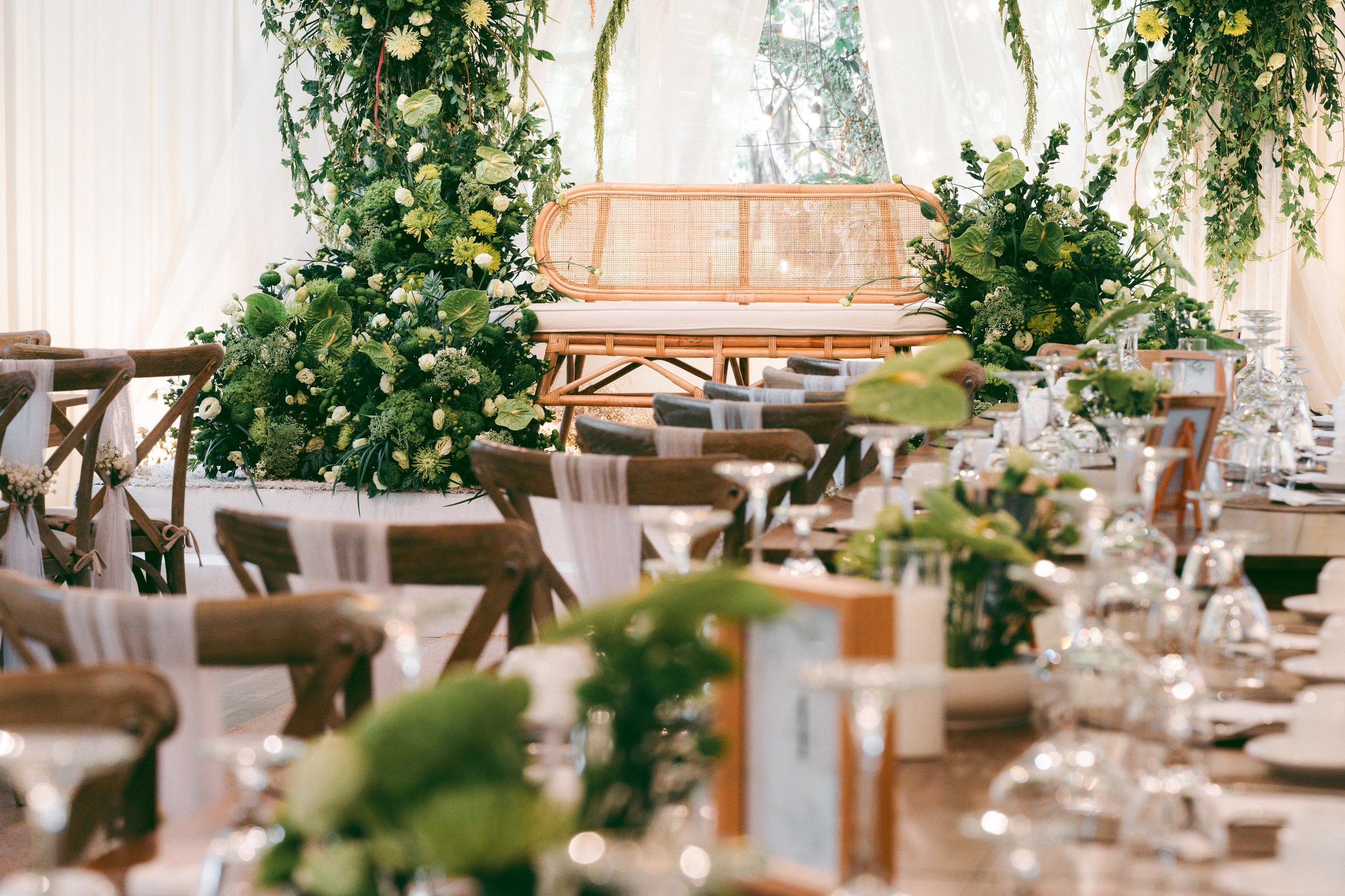 Outdoor wedding reception setup with a wicker loveseat on a decorated stage and a long table with greenery centerpieces in a bright, airy setting.